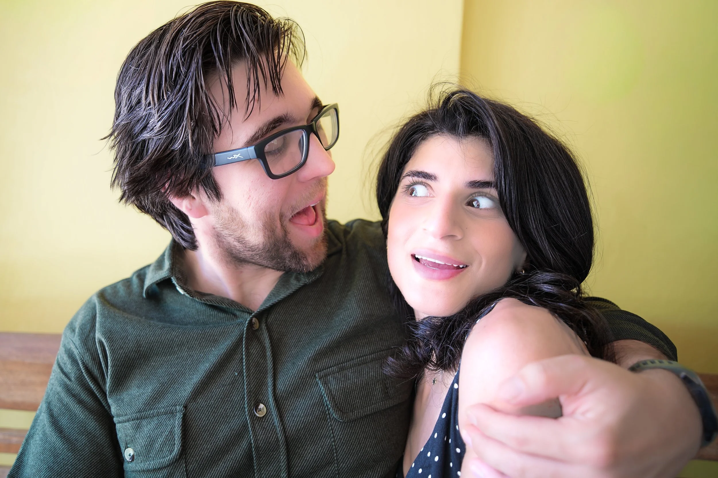 A man with dark, wavy hair, glasses, and a beard taking a selfie with a woman who has long, dark hair and light skin. They are indoors near a yellow wall, smiling and close together.