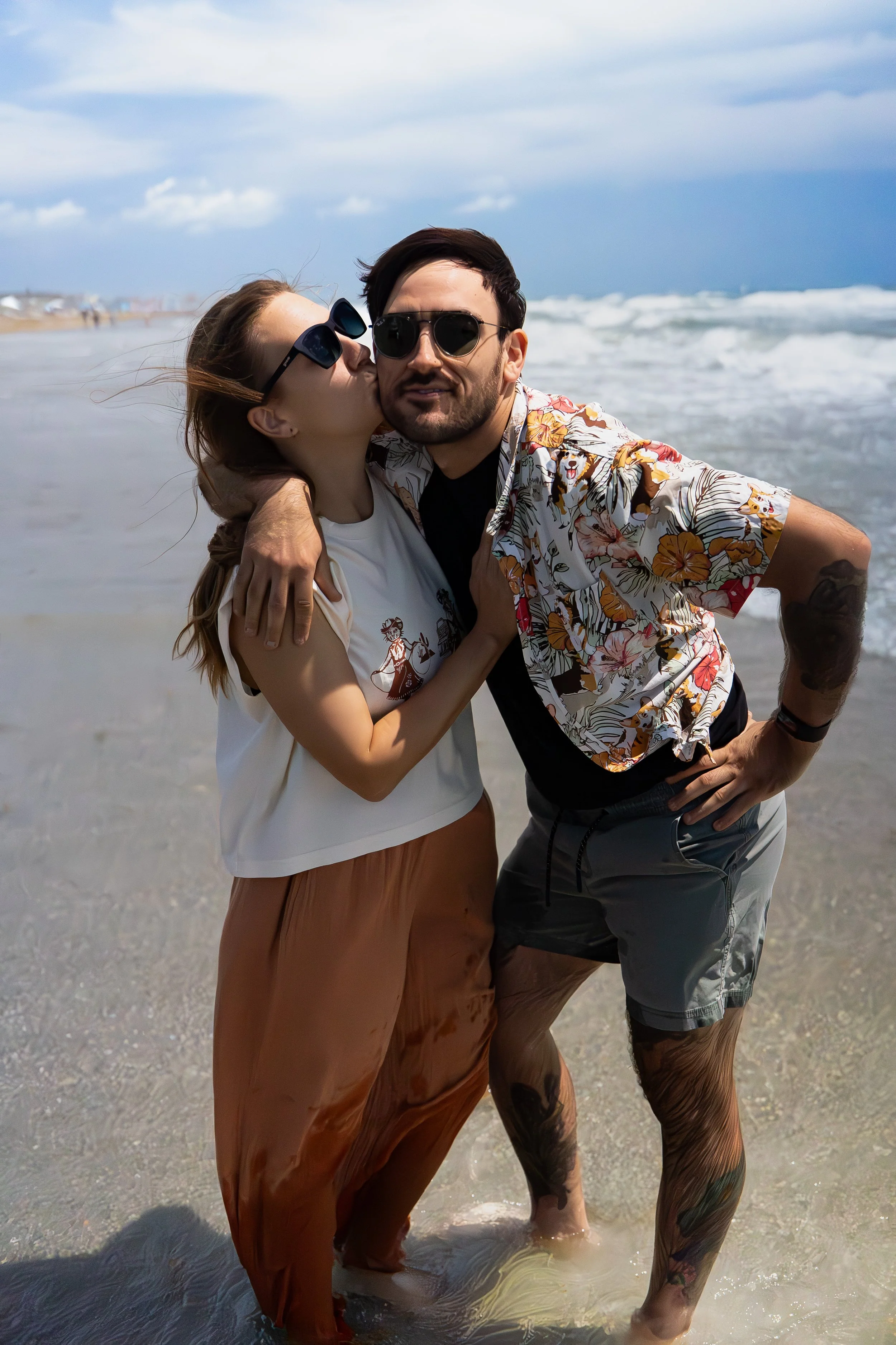 A couple standing in the ocean on the beach, with the woman kissing the man on the cheek. They are both wearing sunglasses and casual summer clothing. The background shows waves and a cloudy sky.