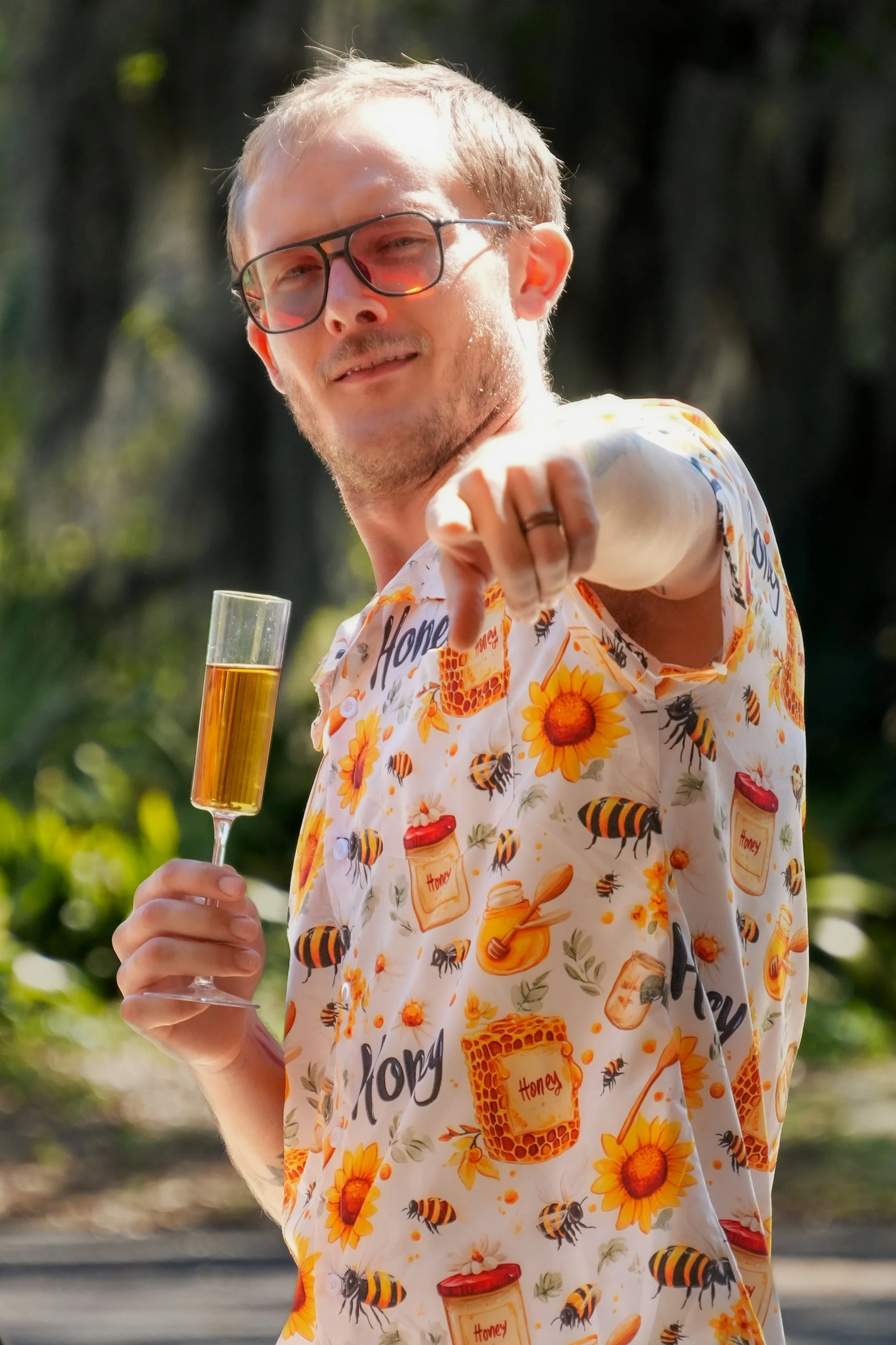 A man with glasses and a beard wearing a short-sleeve button-up shirt with honey and bee patterns, holding a glass of champagne and pointing towards the camera outdoors.