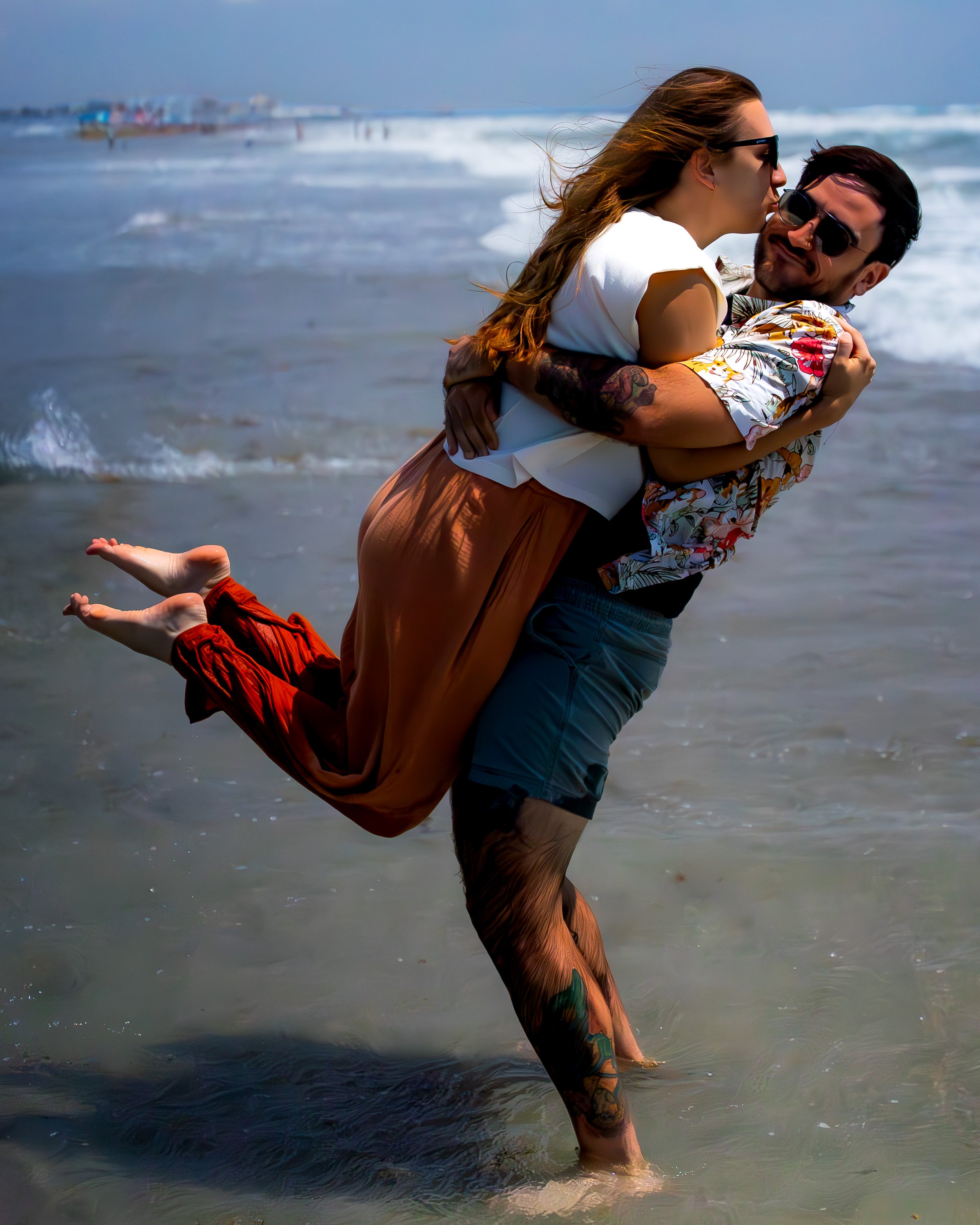 Couple at the beach, with the woman riding on the man's back, both wearing sunglasses, with waves and a pier in the background.
