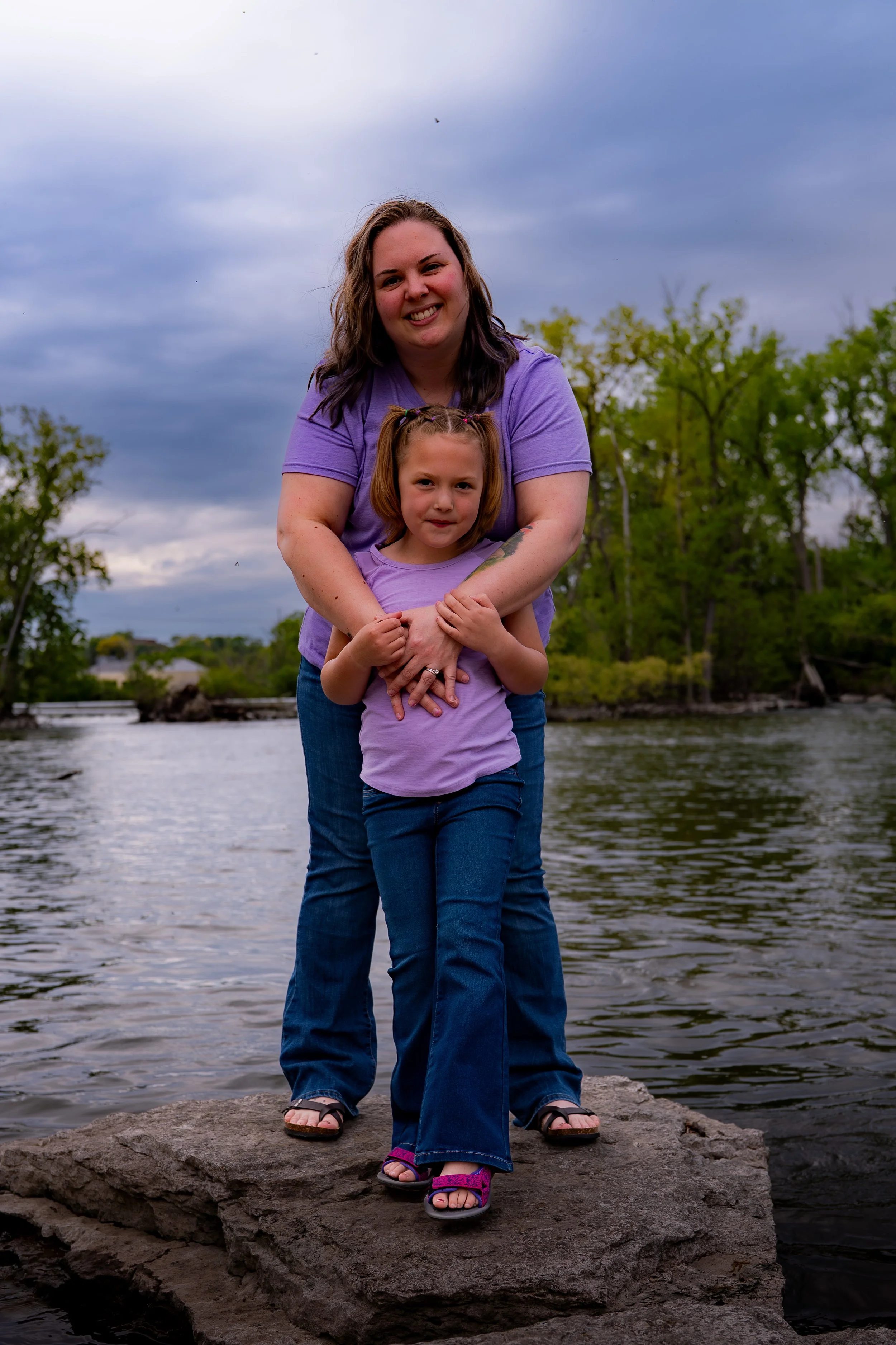 A woman and a young girl standing on a rock near a body of water with trees in the background. The woman is smiling and wearing a purple shirt, while the girl in front of her has pigtails and is also smiling, wearing a purple top and jeans.