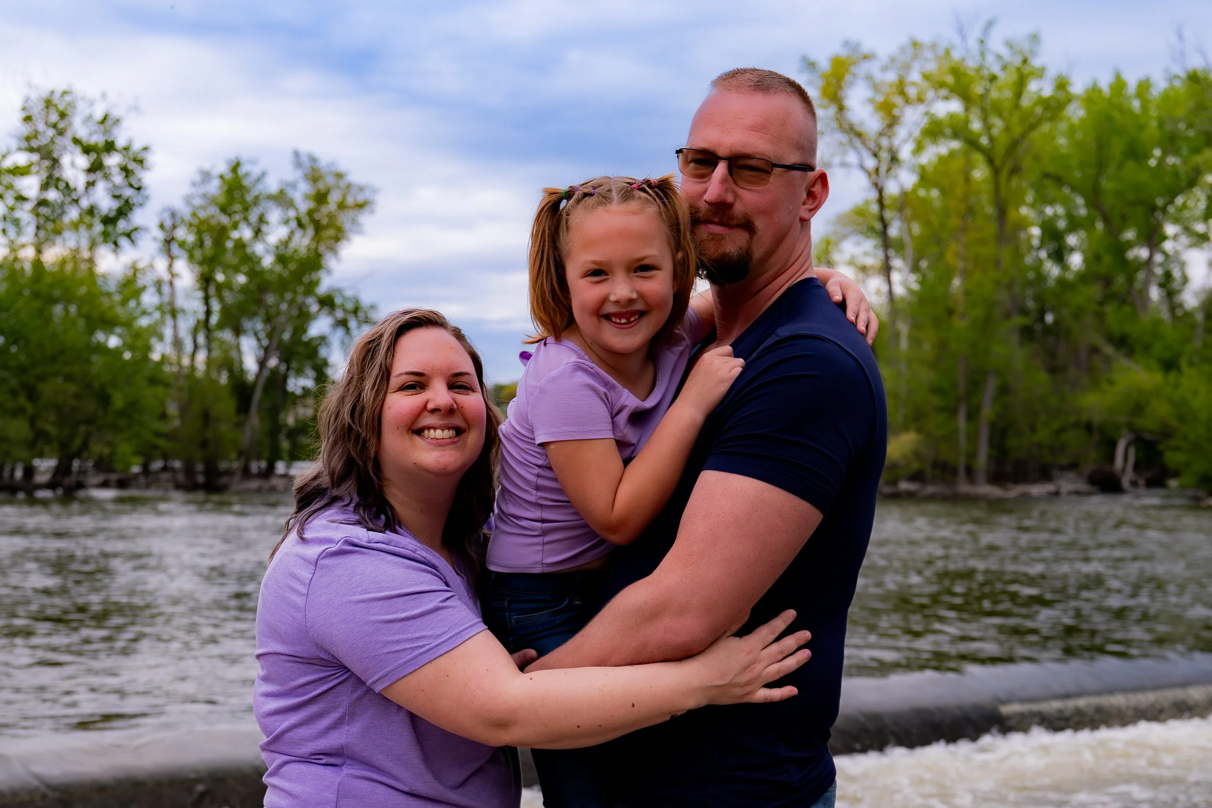 A family of three, with two women and a man, standing near the water with trees in the background. They are smiling and hugging, with the man holding a young girl between them.