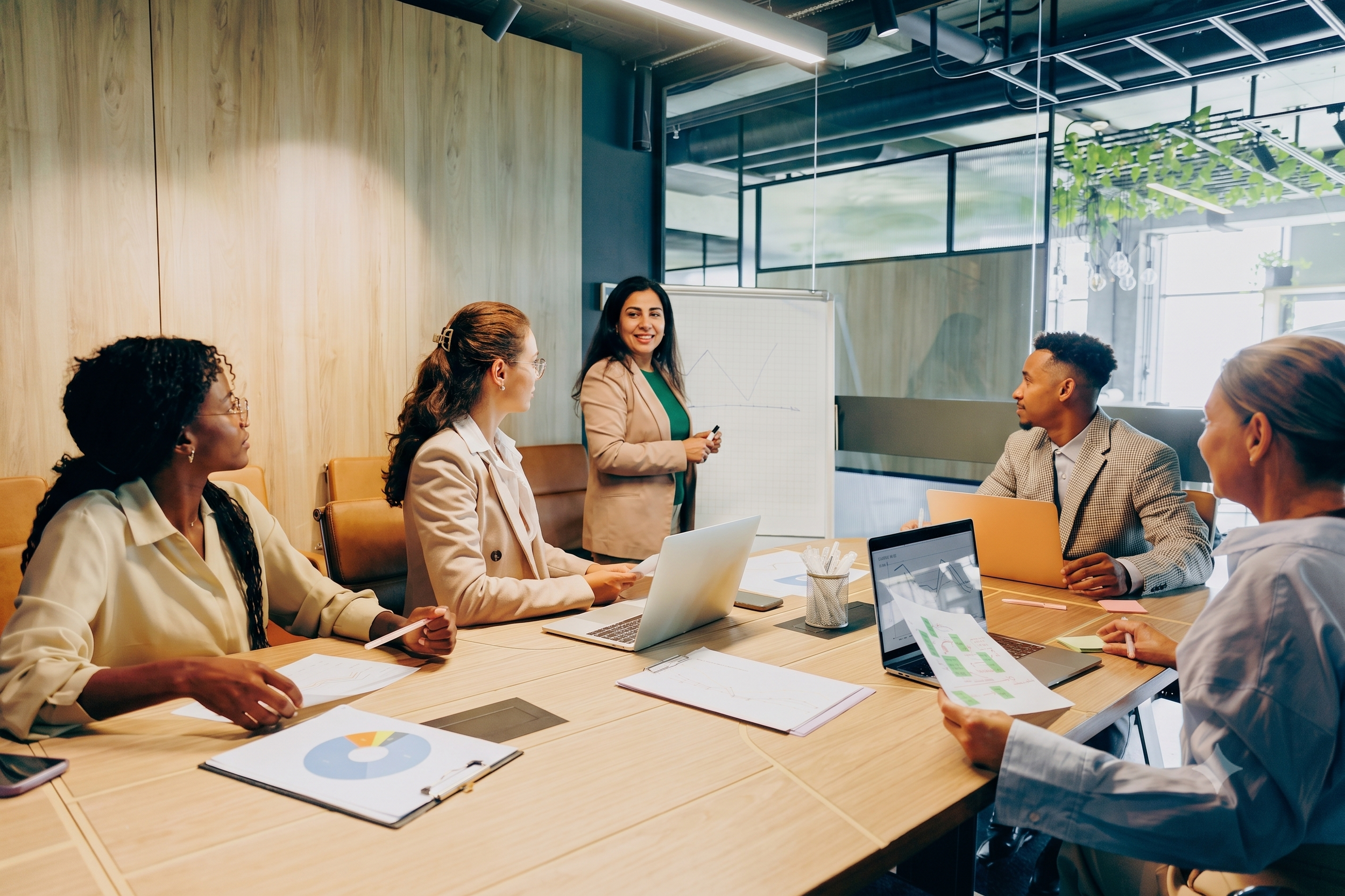 A diverse group of five professionals in business attire participating in a team meeting in a modern conference room with laptops, documents, and presentation materials on the table.
