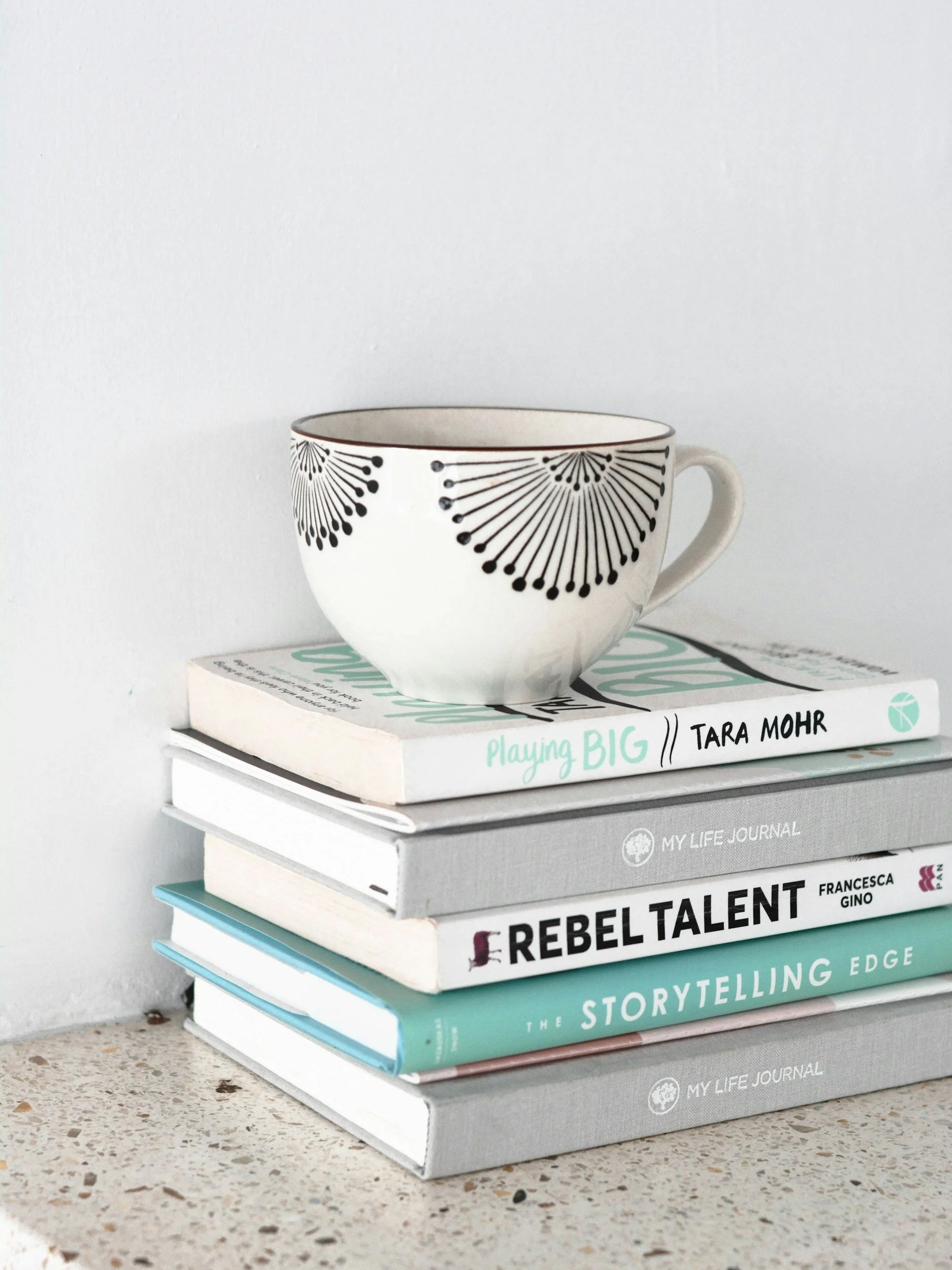 A stack of various books with a white patterned mug on top, placed on a speckled countertop against a plain wall.