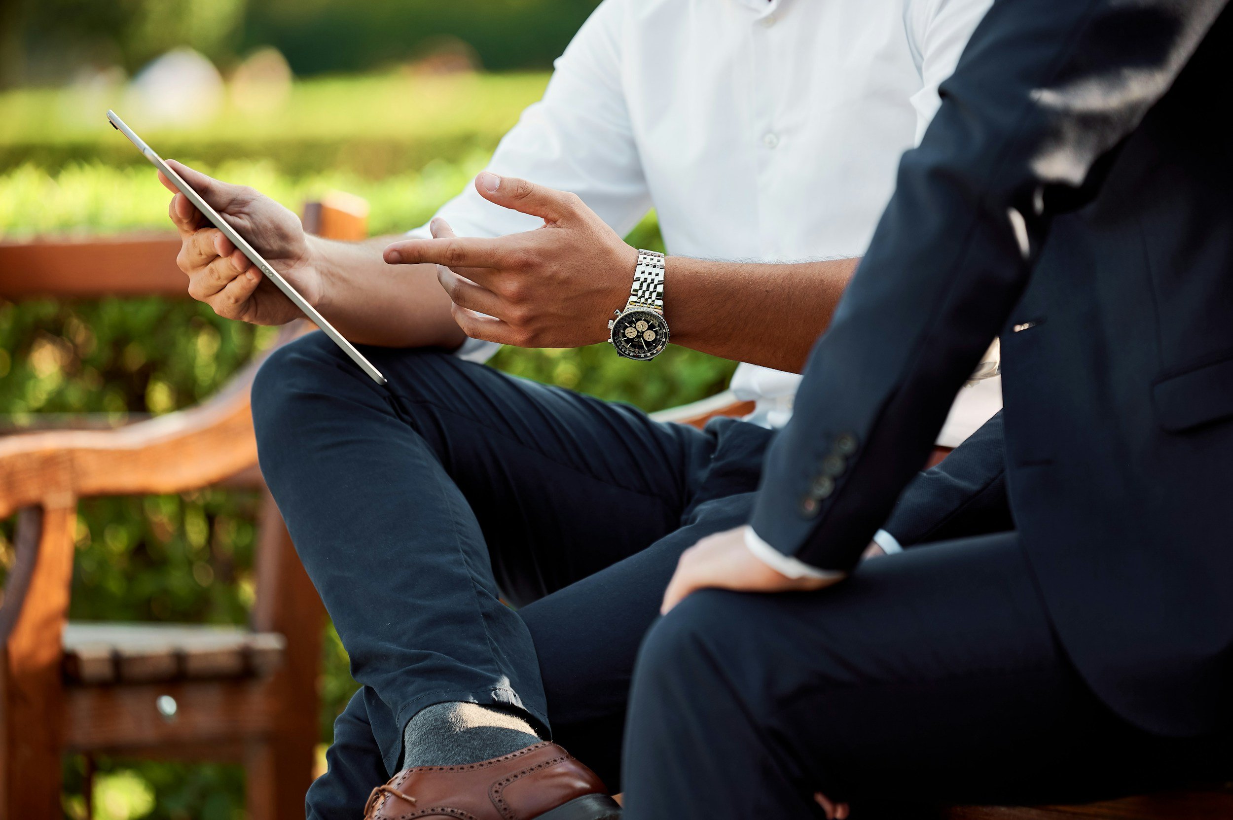 Two men sitting outdoors on a park bench, one wearing a white shirt and a silver watch, holding a tablet, the other wearing a dark blazer and white shirt, in a green park setting.