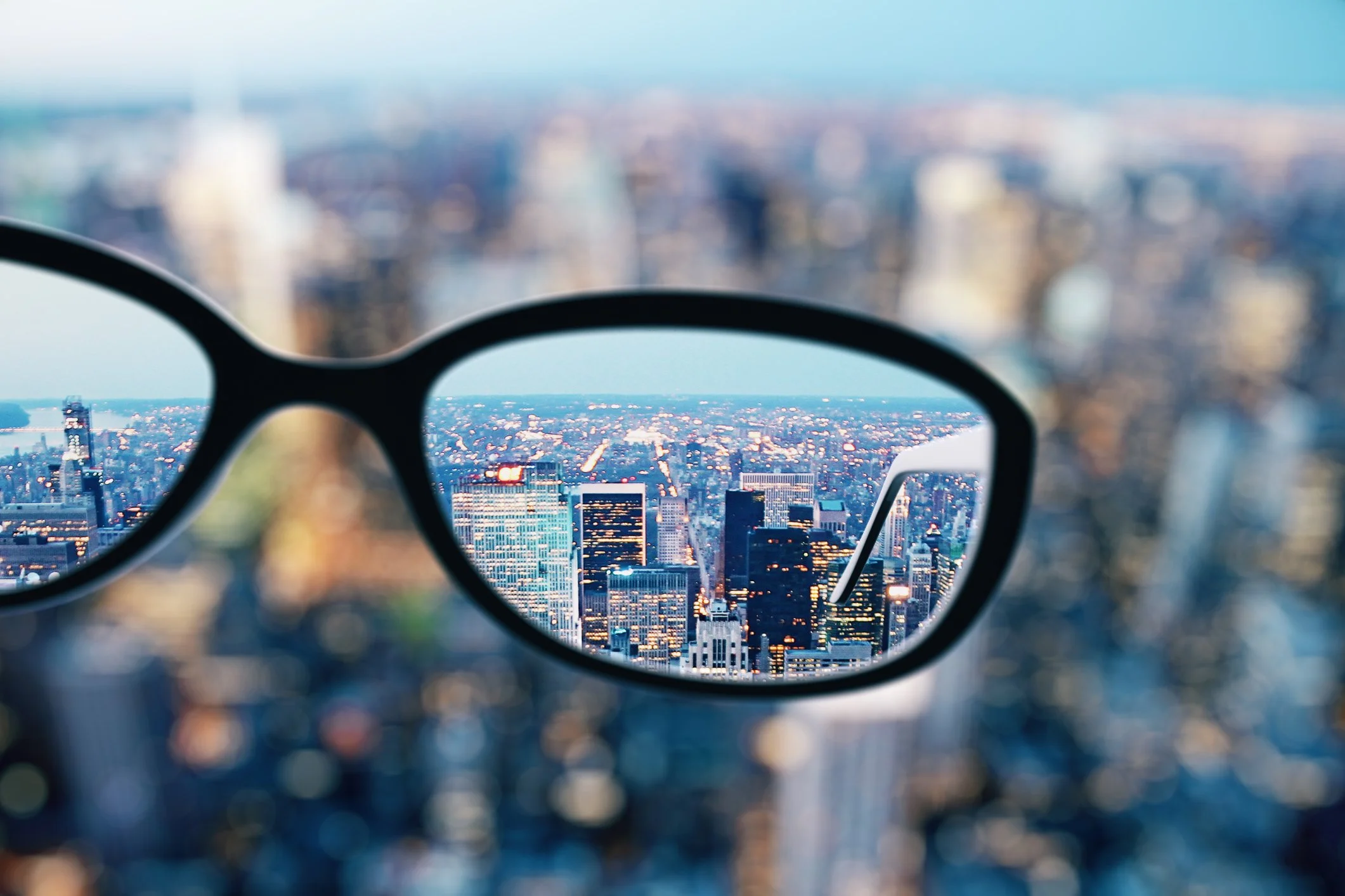 A pair of glasses with a city skyline view of skyscrapers and buildings, blurred city, and blue sky in the background.