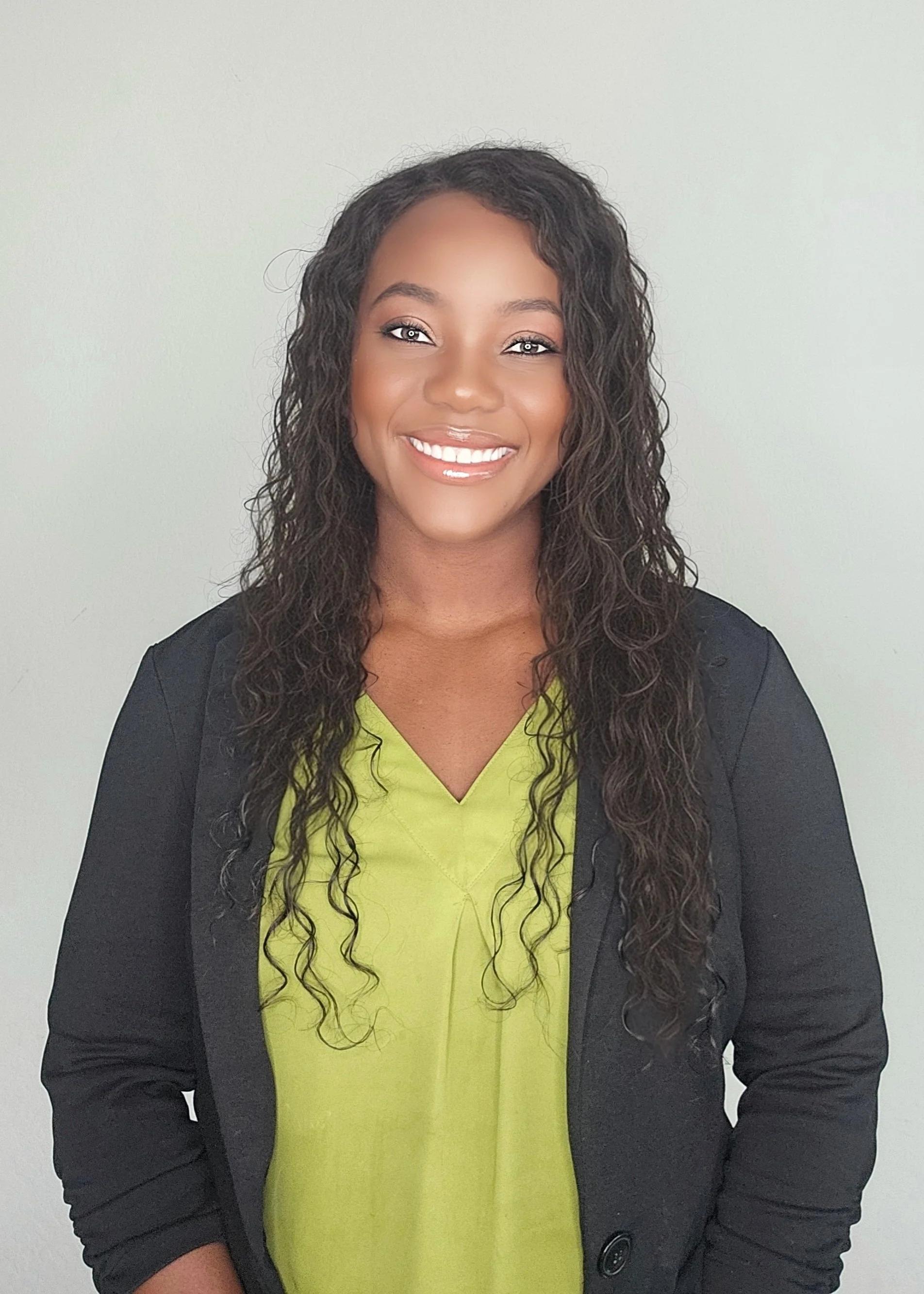 A woman with long, curly dark hair, wearing a black blazer over a lime green top, smiling at the camera against a plain white background.