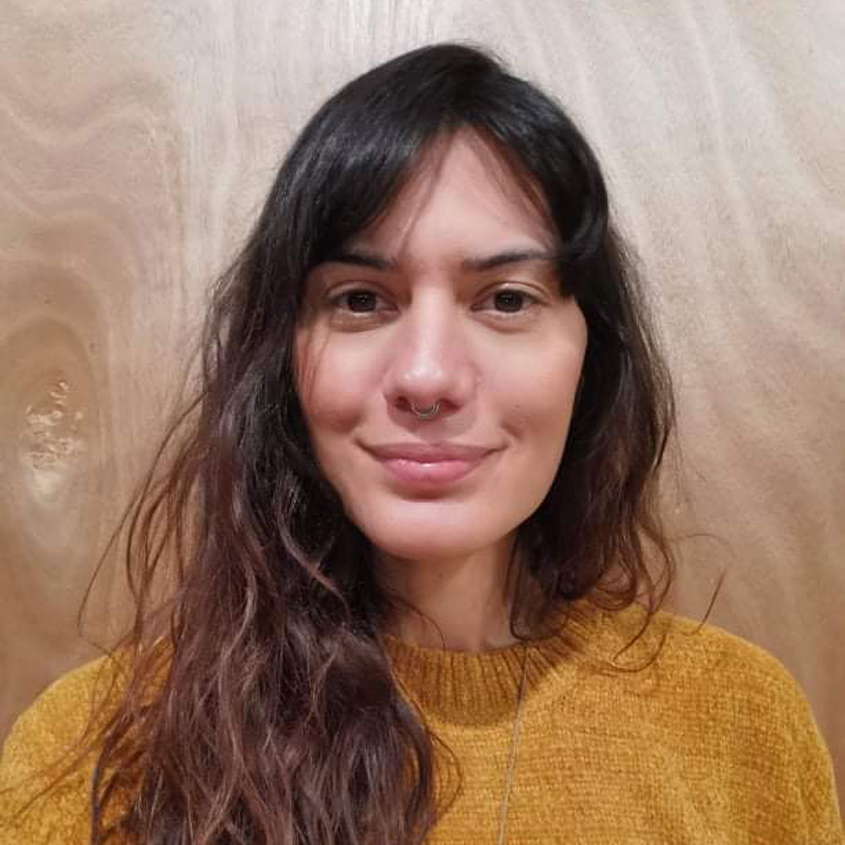 A woman with shoulder-length dark wavy hair, a septum piercing, and wearing a mustard yellow sweater, standing in front of a wooden background.