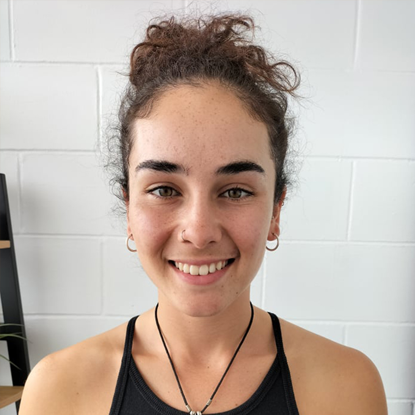 A young woman with curly brown hair in a bun, wearing a black tank top, earrings, a nose ring, and a necklace, smiling in front of a white brick wall.