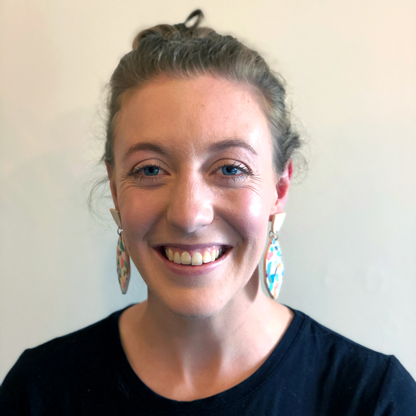 A smiling woman with light skin, blue eyes, and light brown hair pulled back, wearing colorful earrings and a black top, standing against a plain light-colored background.