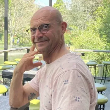 A man with glasses and a bald head, smiling outdoors with trees and empty yellow and gray seating behind him.