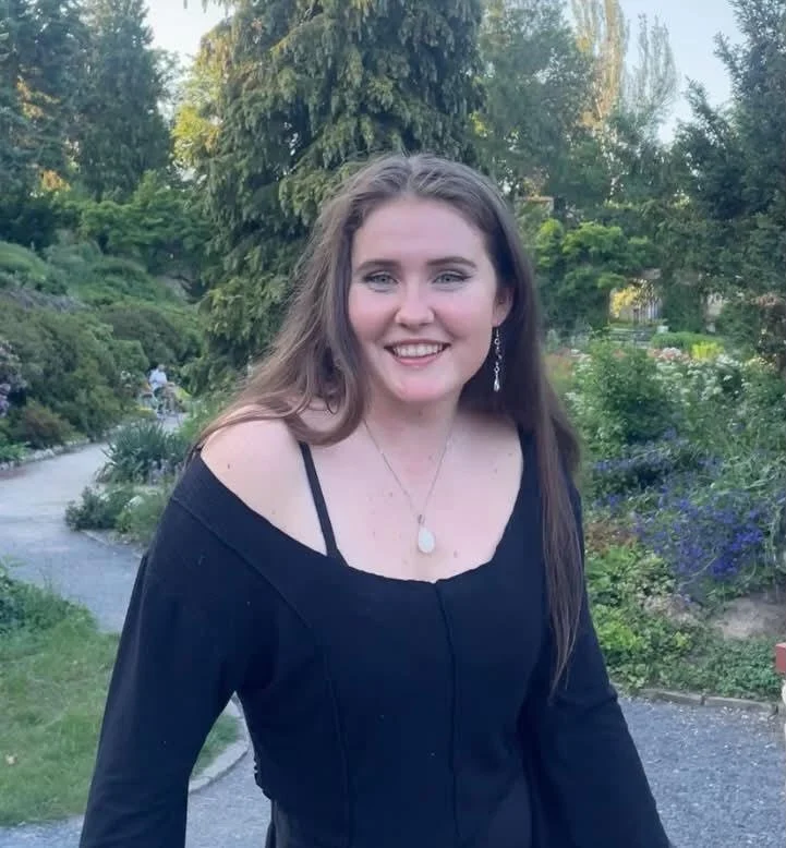 A young woman with long brown hair smiling outdoors in a garden with trees and flowers.