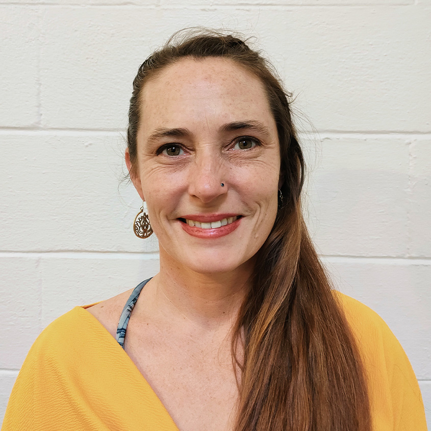 A woman with long brown hair, smiling, wearing a yellow top, silver earrings, and a nose piercing, standing against a white brick wall.