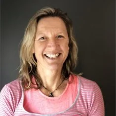 Smiling woman with shoulder-length wavy hair wearing a pink top against a dark background.