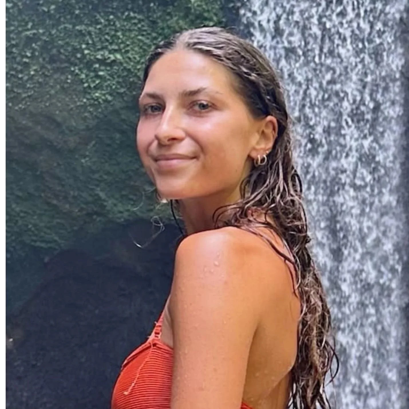 A woman with long wet hair, smiling, standing in front of a waterfall, wearing an orange top.