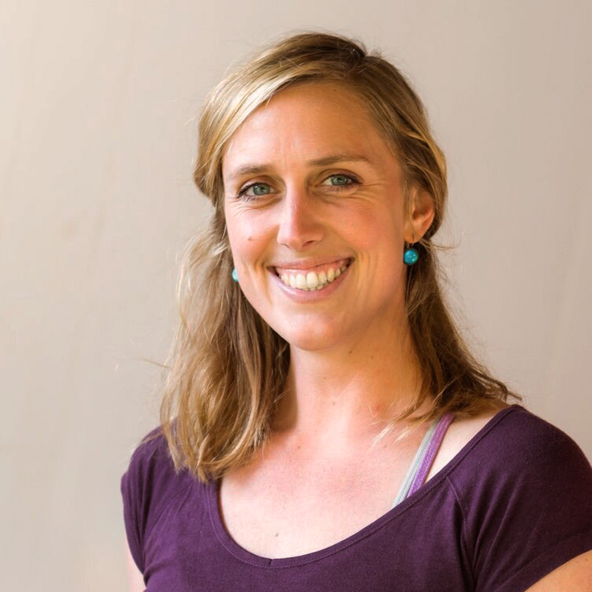 A smiling woman with blonde hair wearing a purple shirt and turquoise earrings.
