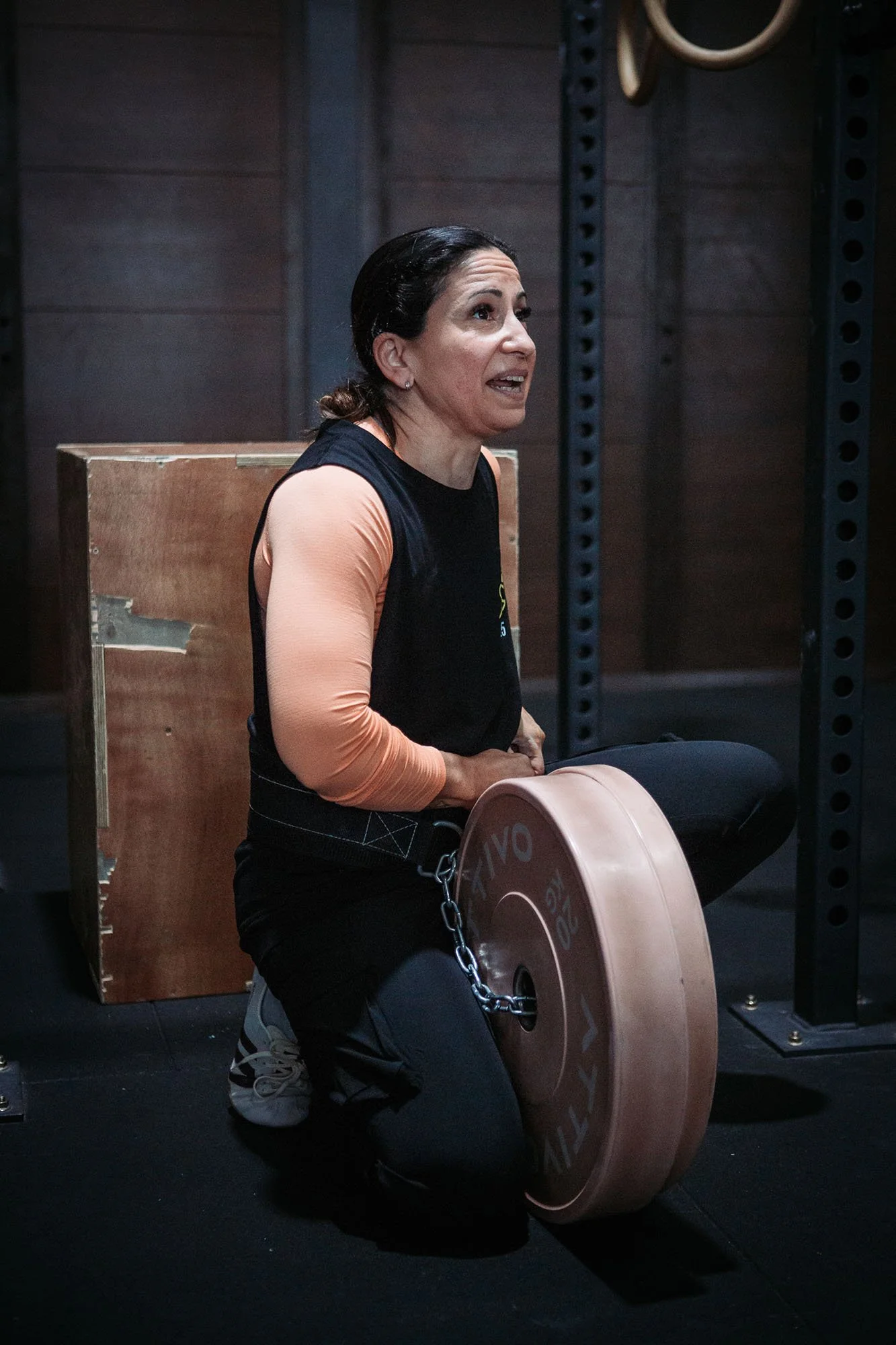 A woman is in a gym, kneeling on one knee, preparing to lift a heavy weight plate. She is wearing a black workout shirt with peach-colored sleeves, black workout pants, and sneakers, and is focusing on her exercise.
