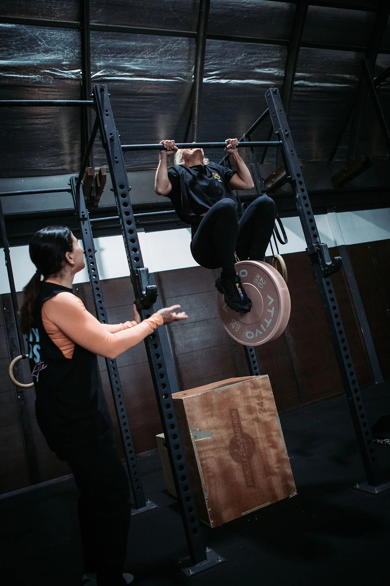 A woman is performing a pull-up exercise on a pull-up bar in a gym, with a trainer standing nearby observing and providing support.