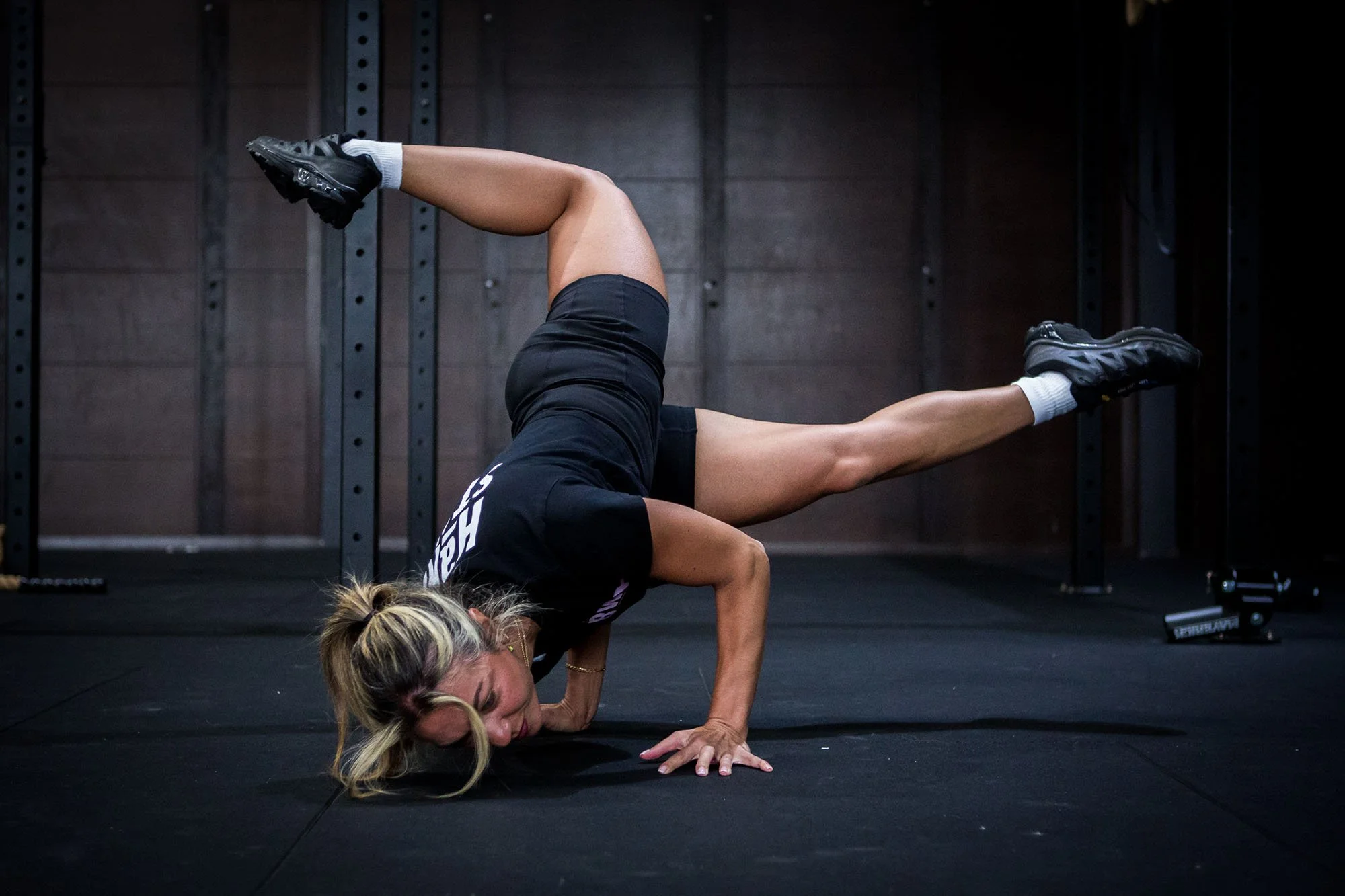 A woman wearing a black shirt, black shorts, and sneakers is performing a handstand split in a gym. She is balancing on her head and hands with one leg extended straight up and the other extended to the side. The gym has a dark interior with black flooring, and metal wall supports are visible behind her.