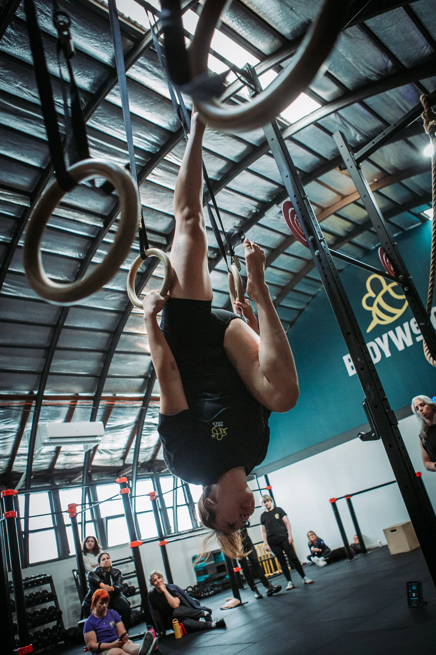 A woman performing an exercise on gymnastic rings in a gym, with a group of people sitting and standing around watching.