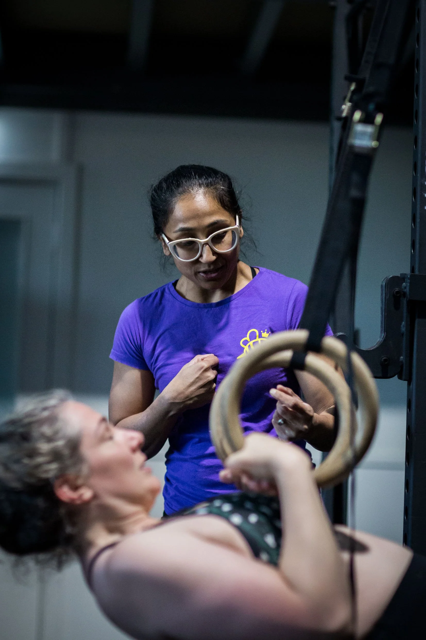Two women, one with dark hair and glasses, and another with blonde hair, are in a gym. The woman with dark hair is wearing a purple shirt and appears to be coaching or guiding the woman with blonde hair during an exercise involving gymnastic rings.