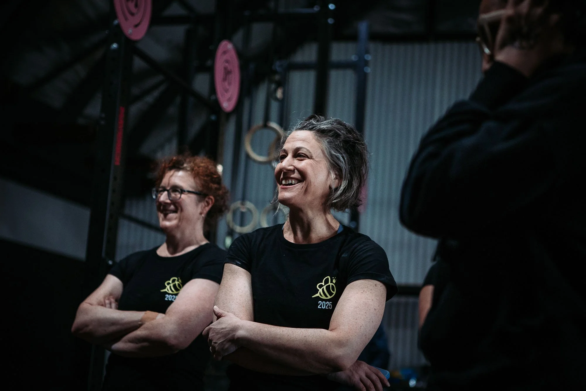 Three women stand in a gym, smiling and smiling with their arms crossed, wearing black shirts with a yellow bee logo printed on them. They are in front of a gym structure with pink and beige gymnastic rings hanging from it.