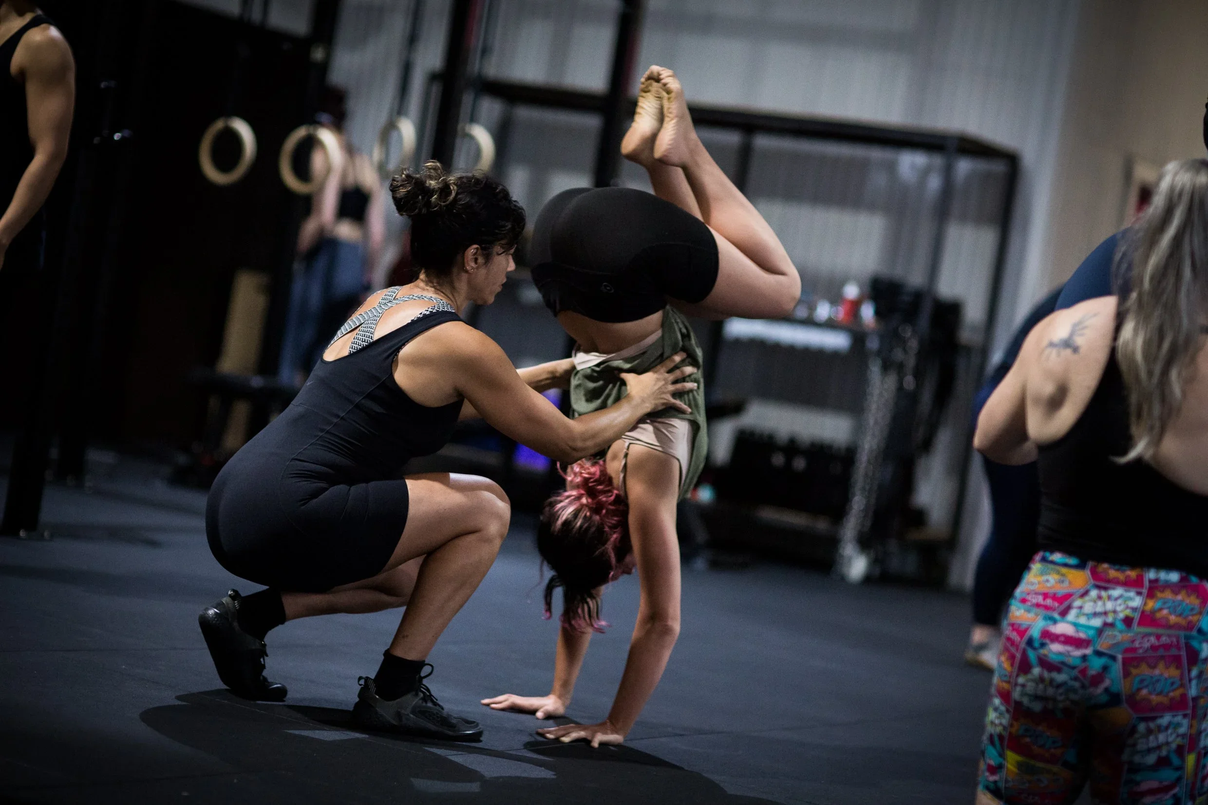 A woman coaching a woman with pink hair do a handstand in a gym, with rings hanging in the background.