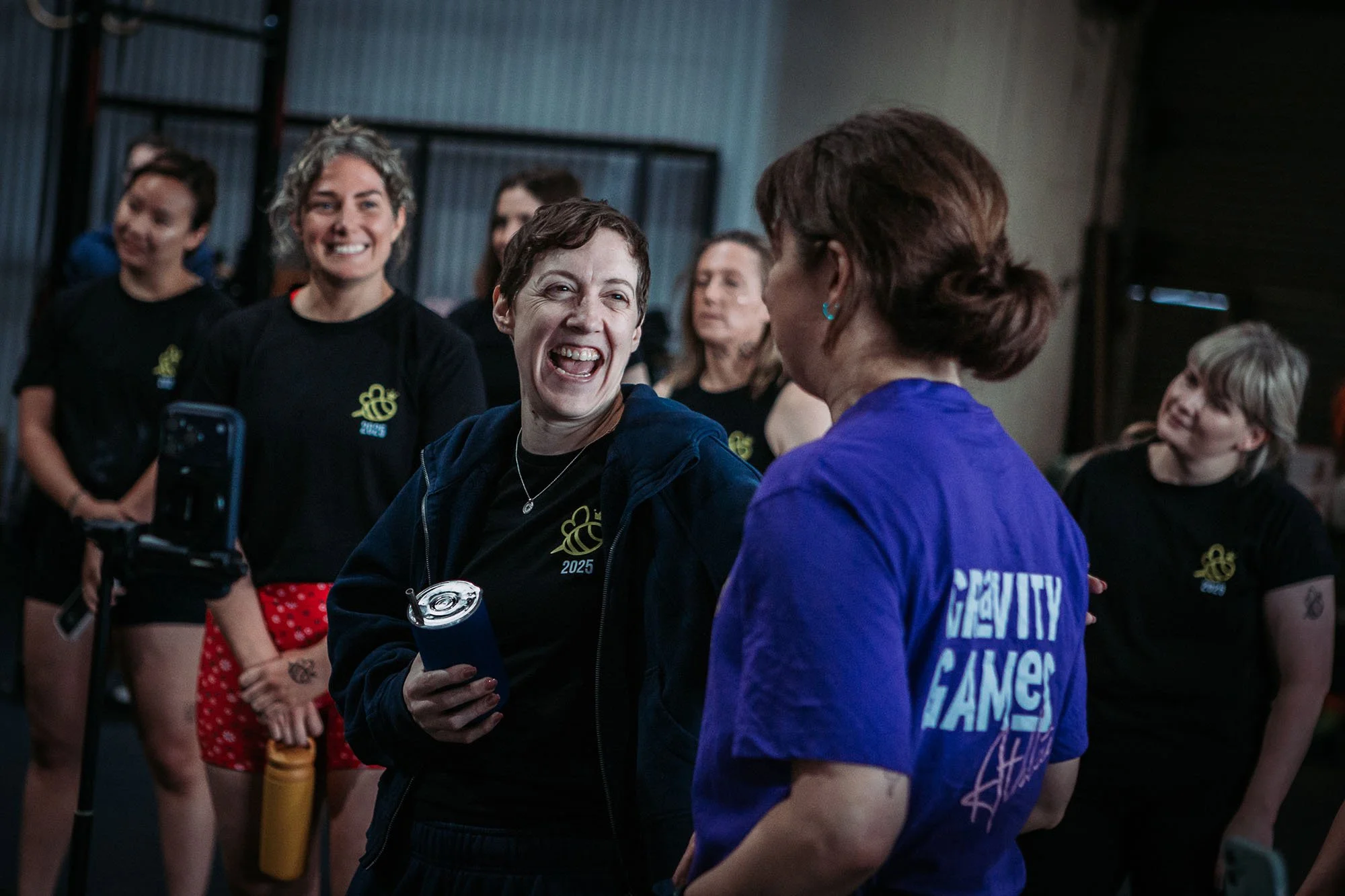 Group of smiling women, some wearing black shirts with a yellow bee logo, gathered in a gym, engaging in conversation and smiling.