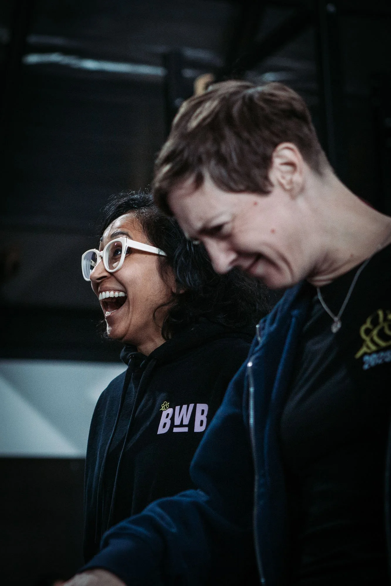Two women, one with dark curly hair and glasses and the other with short brown hair, are smiling and laughing together indoors. Both are wearing black hoodies, and the woman with glasses has a logo on her hoodie.