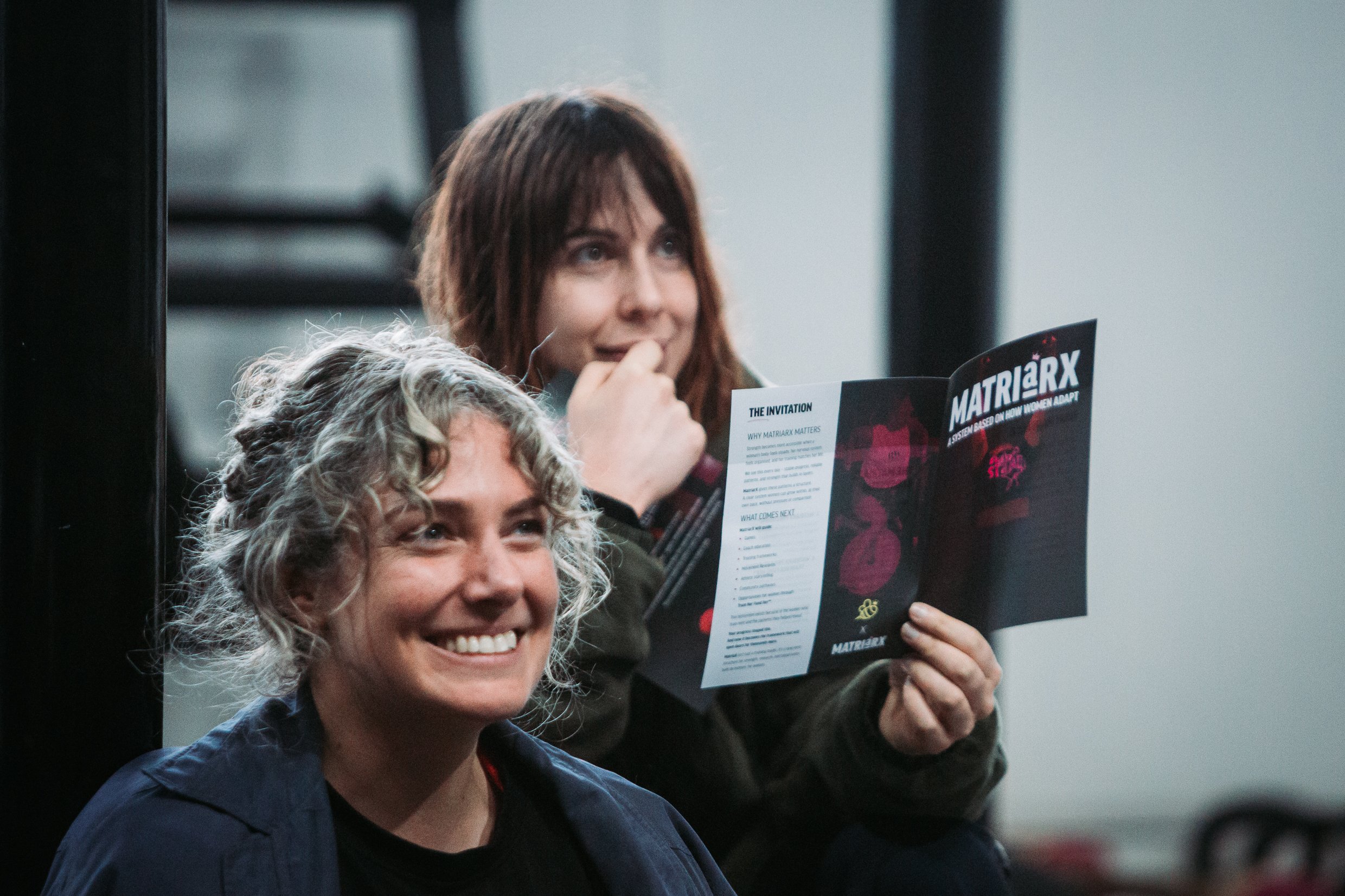Two women smiling; the woman in the foreground has gray, curly hair and is wearing a blue jacket; the woman in the background has brown hair and is holding a flyer that reads 'MATRIARX®'.