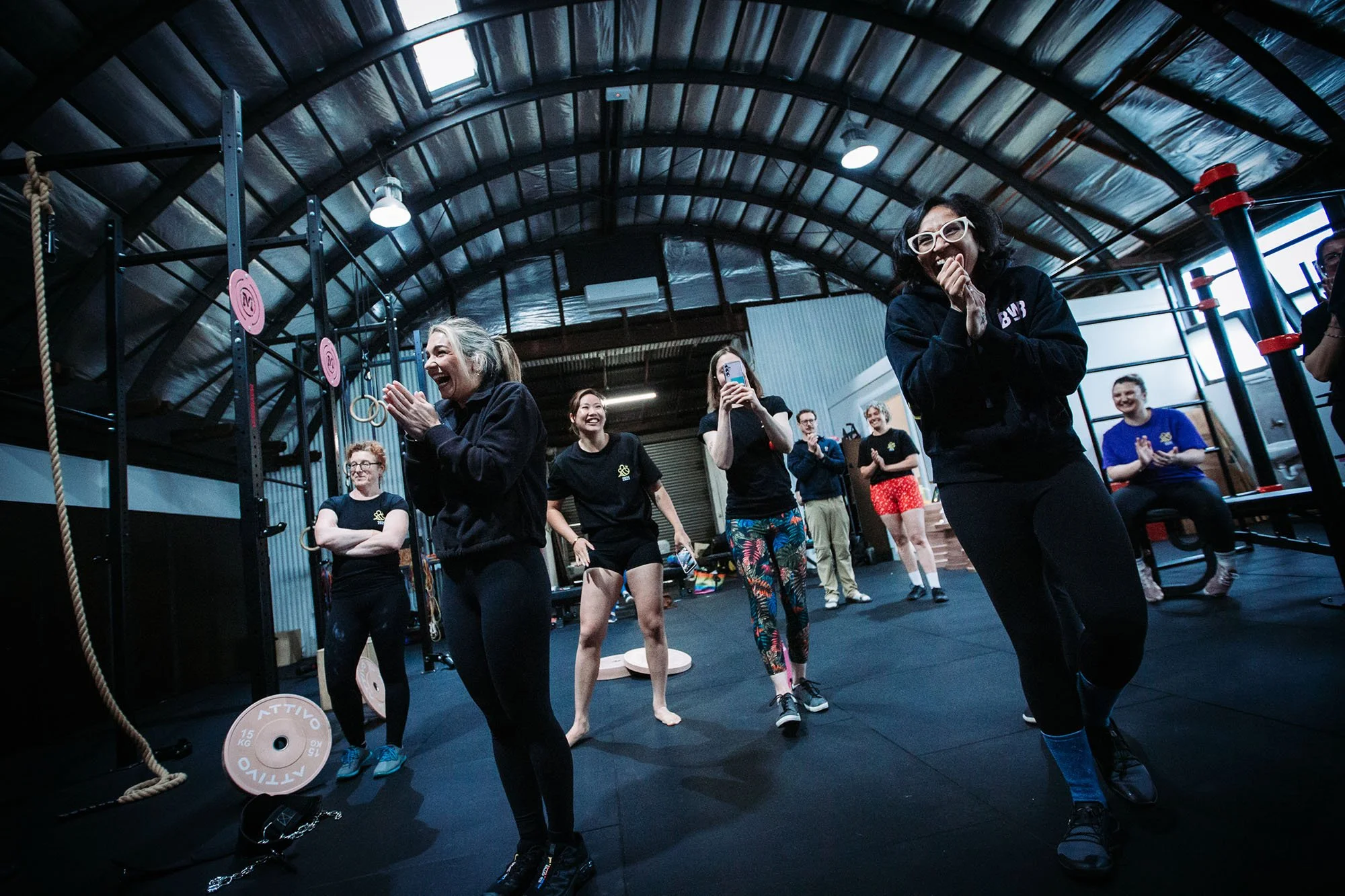 Women socializing in a gym or fitness class setting, with exercise equipment and weights in the background.