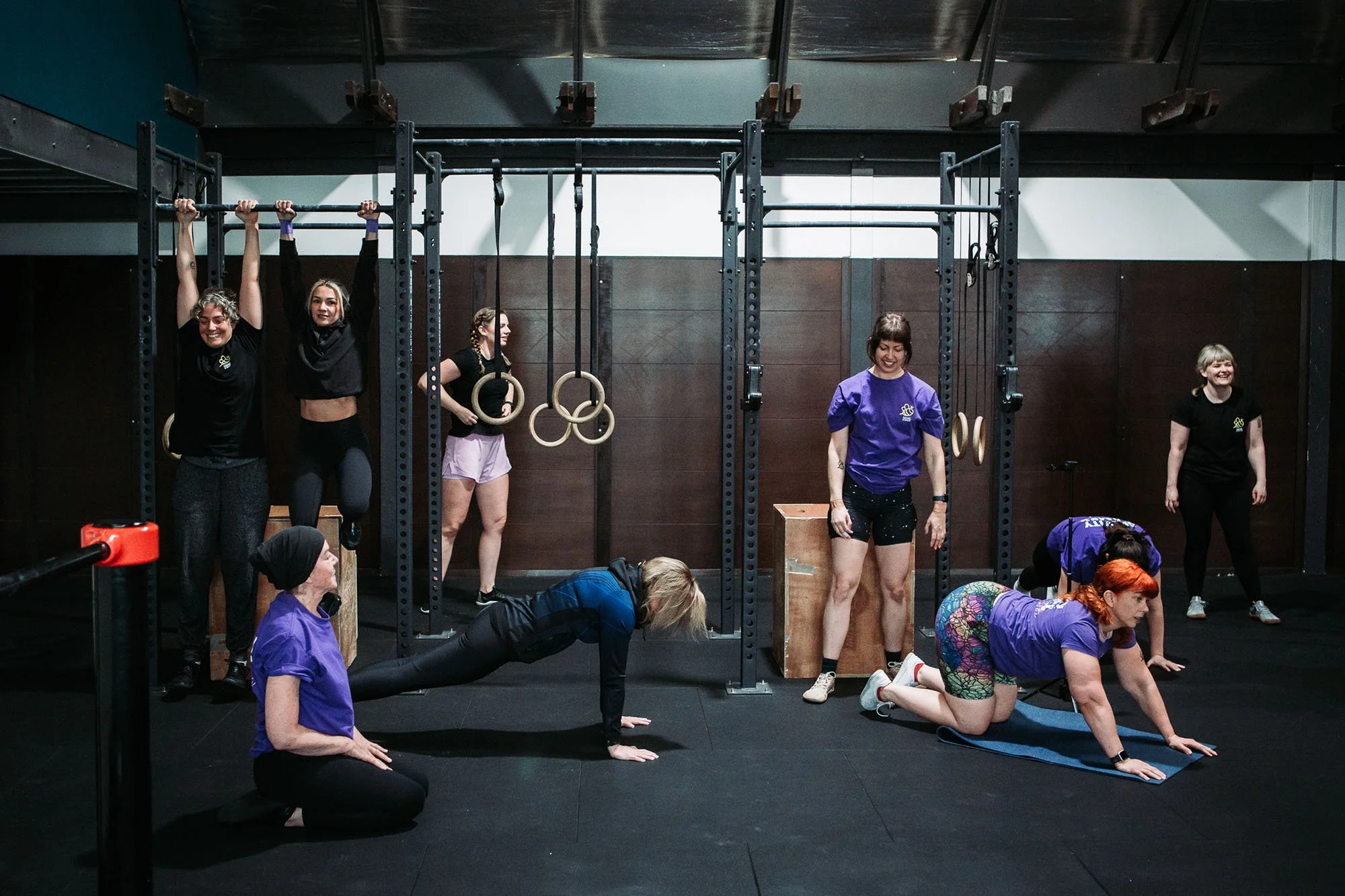 A group of people participating in a fitness class in a gym, with some performing exercises on mats and others using gym equipment, smiling and enjoying the workout.