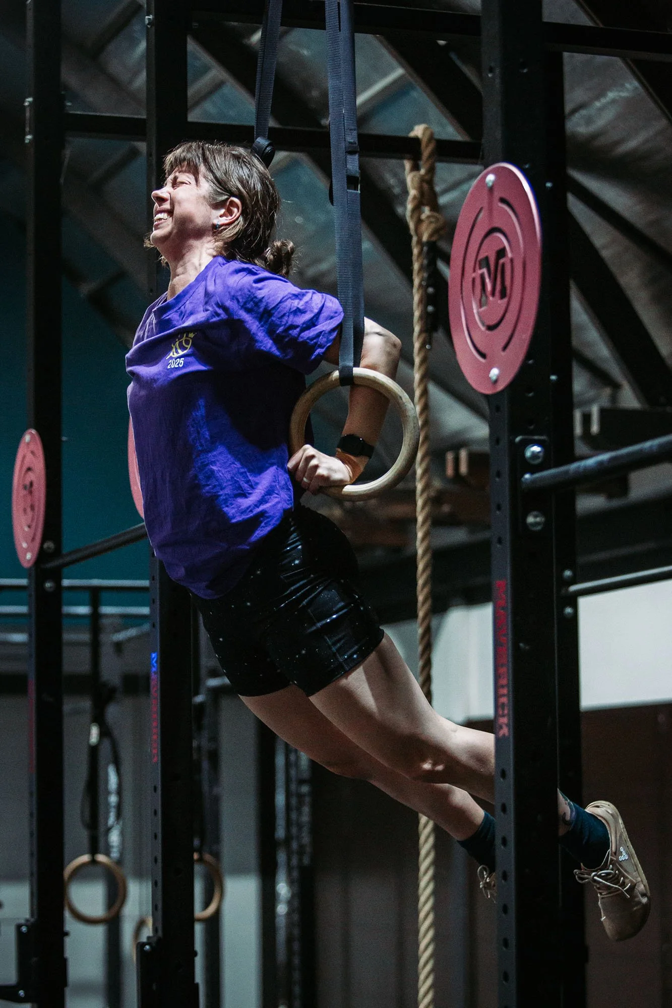 A woman with short brown hair, wearing a purple athletic shirt and black shorts, is performing a hanging leg raise exercise on gymnastic rings in a gym.