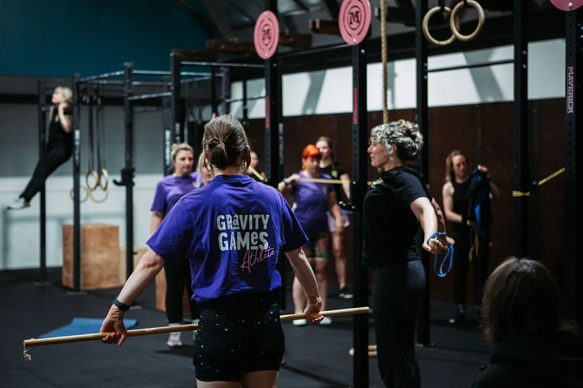Group of women participating in a fitness class at Gravity Games, holding resistance bands and a wooden stick in a gym with workout equipment.