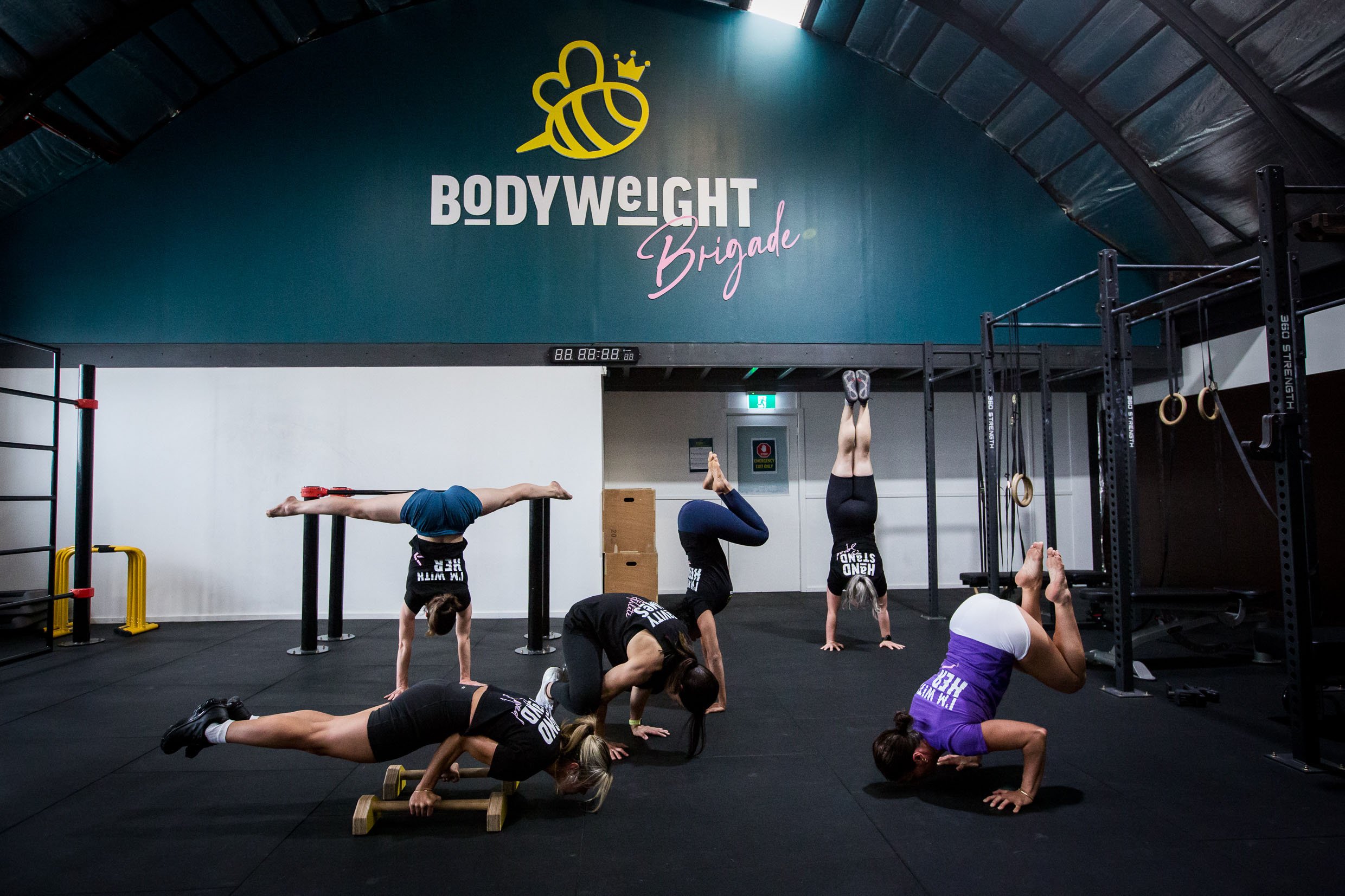 People practicing arm balances in a gym with a large blue wall featuring a bee logo and the words 'Bodyweight Brigade' at the top.