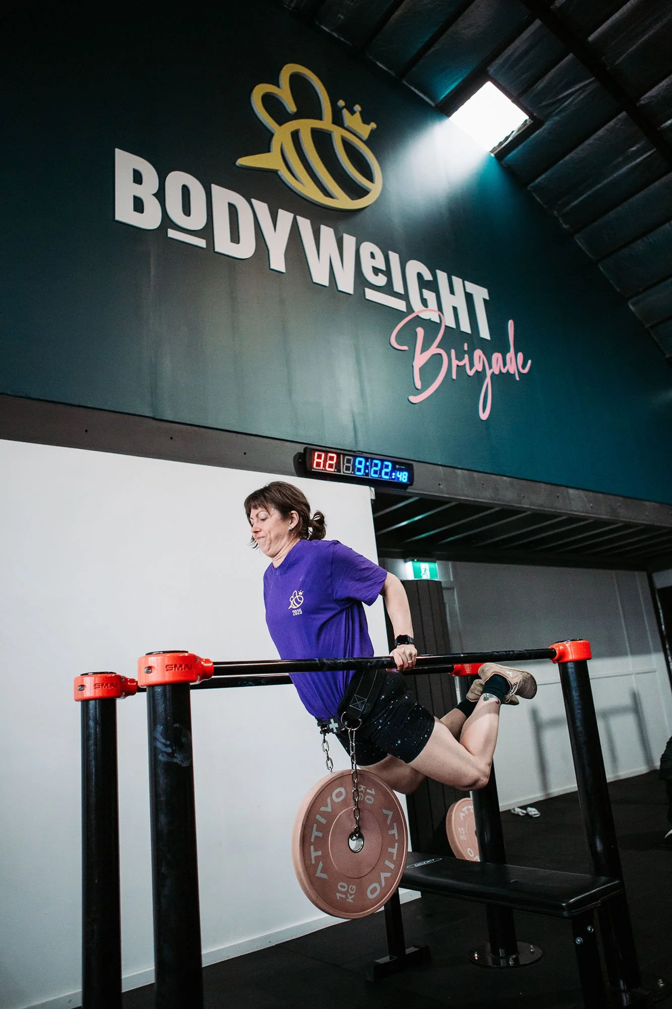 A woman performing a triceps dip exercise on parallel bars in a gym. She is wearing a purple shirt, black shorts, and tan shoes. There is a large sign on the wall with a bee logo and the text 'BODYWEIGHT Brigade'. 