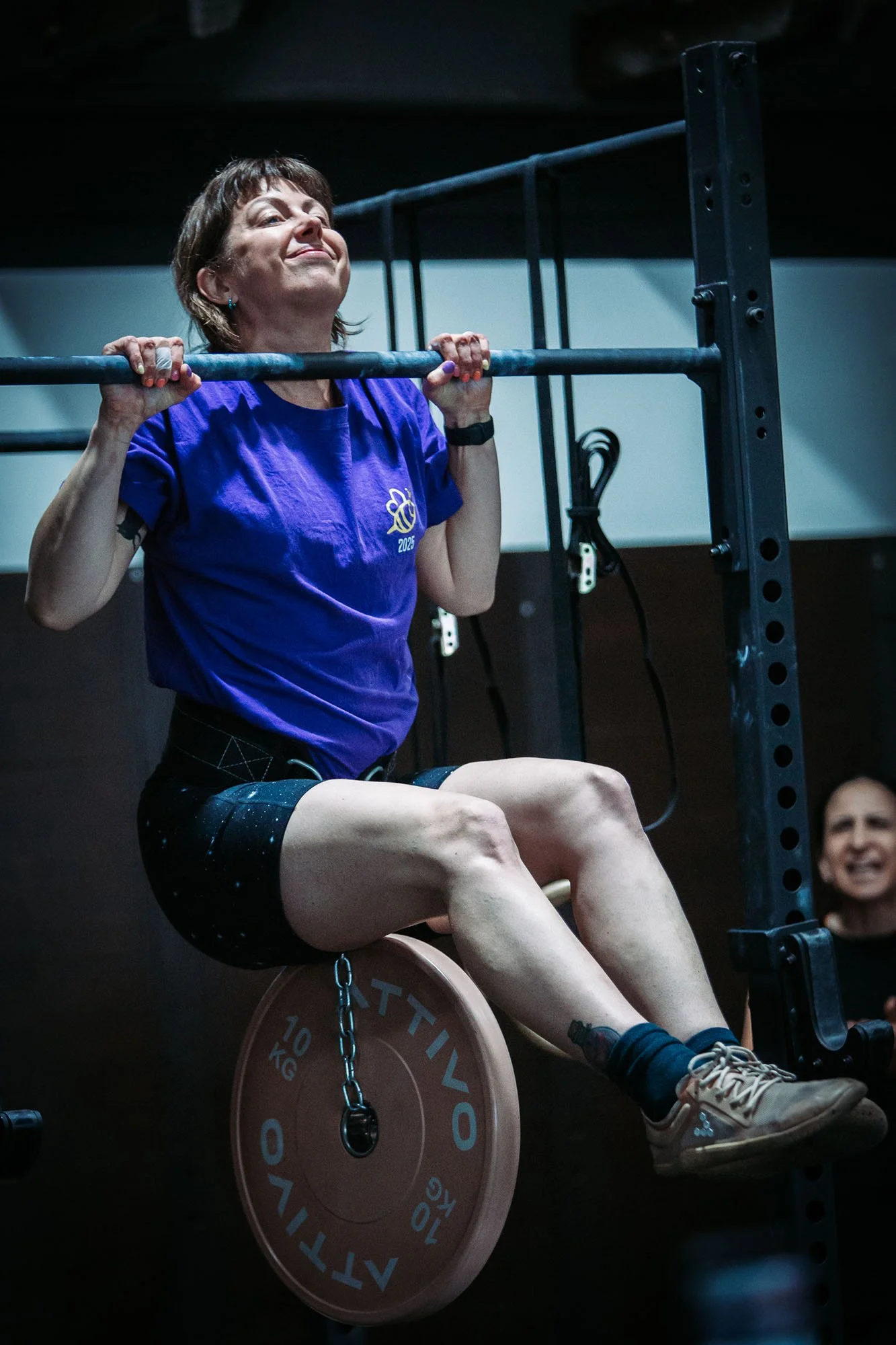 A woman in a purple t-shirt performing a pull-up exercise using a bar, in a gym or fitness setting.