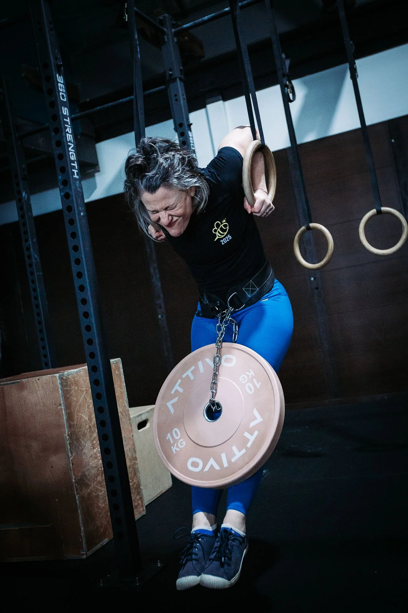 Woman with curly gray hair in black shirt and blue pants performing a ring exercise with gymnastic rings, in a gym setting.