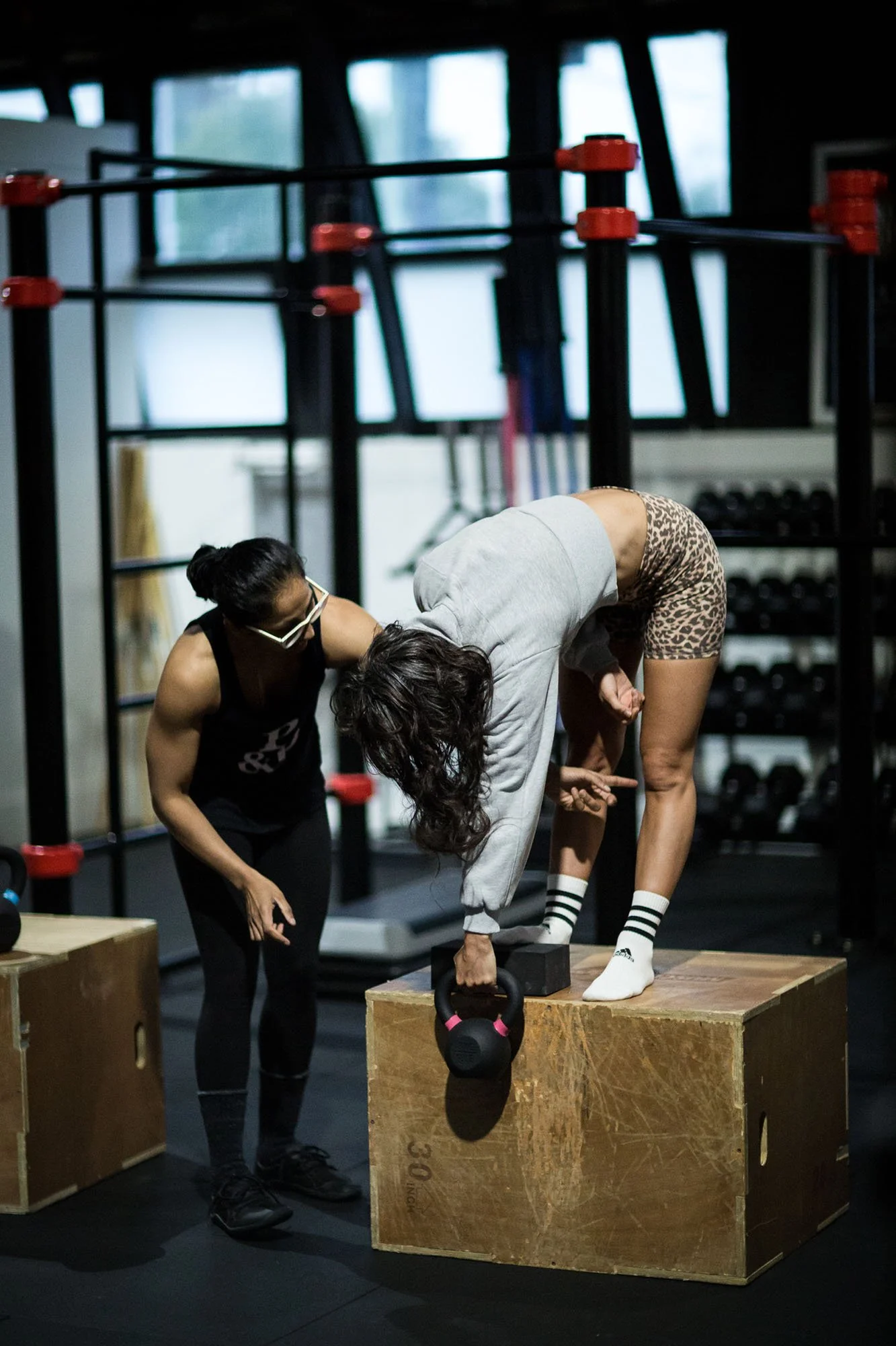 two women in a gym, one is standing on a box performing an exercise with a weight while the other coaches her