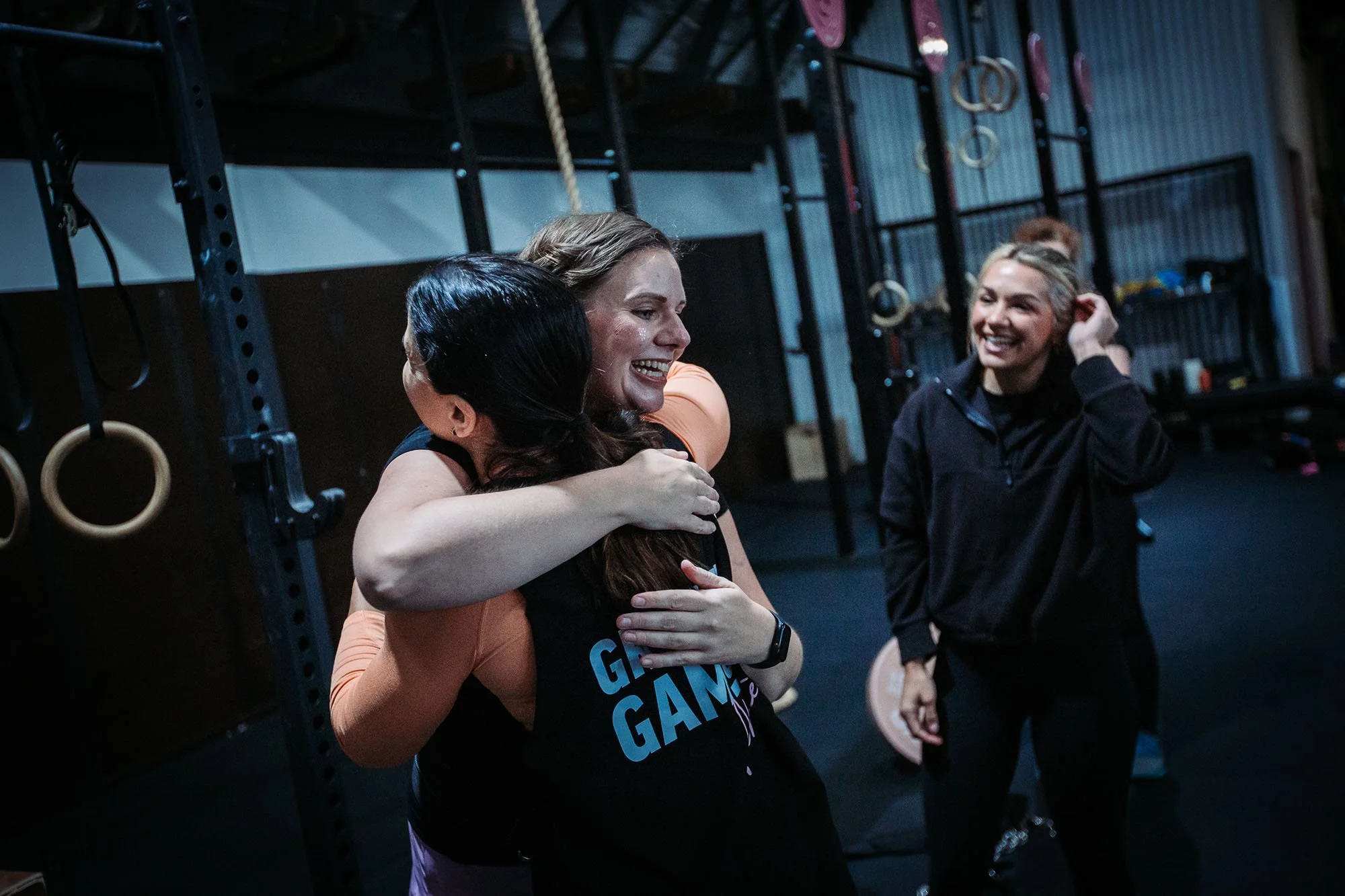 Two women hugging and smiling in a gym, with gymnastic rings and ropes in the background.