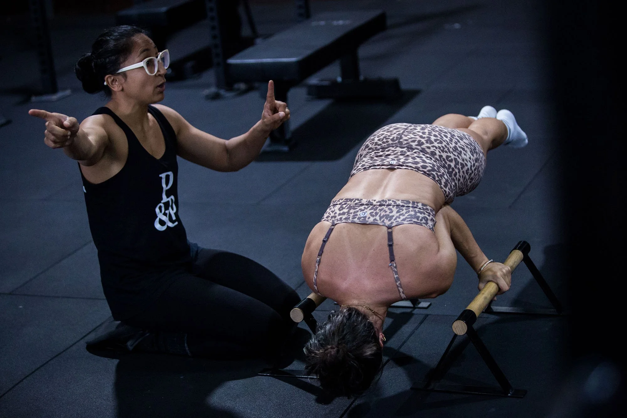 A woman in leopard print workout clothes doing an exercise on a bench while a fitness instructor instructs her in a gym.