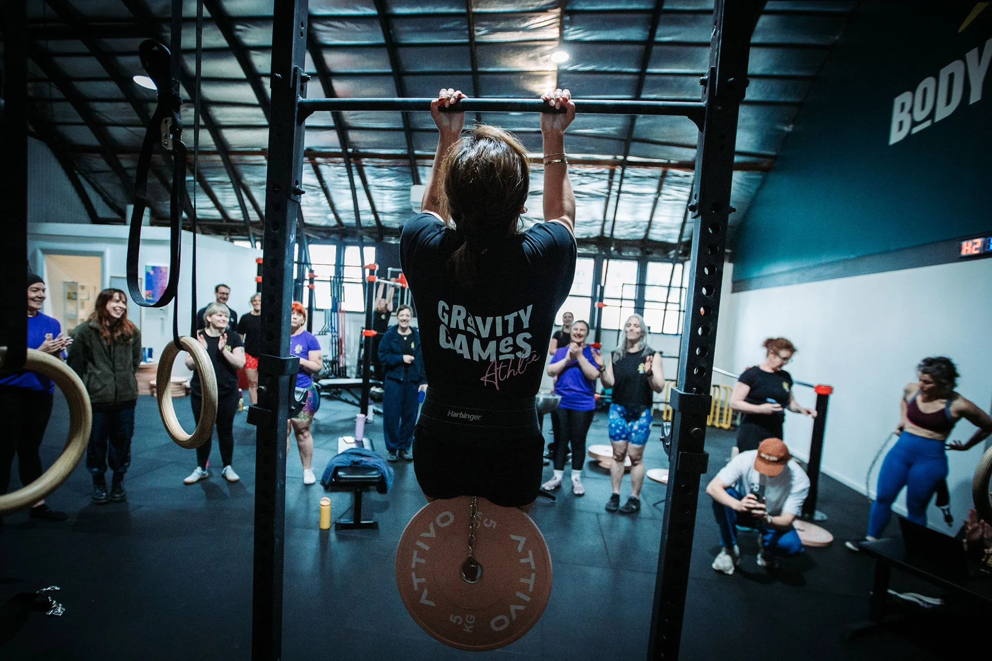 Woman doing pull-up exercises on a pull-up bar in a gym surrounded by a group of people watching and cheering.