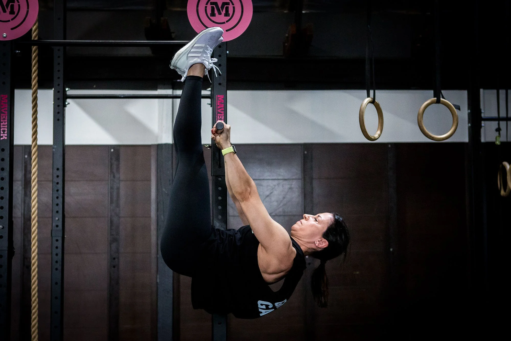 Woman performing a hanging leg raise exercise in a gym, surrounded by gym equipment like rings.