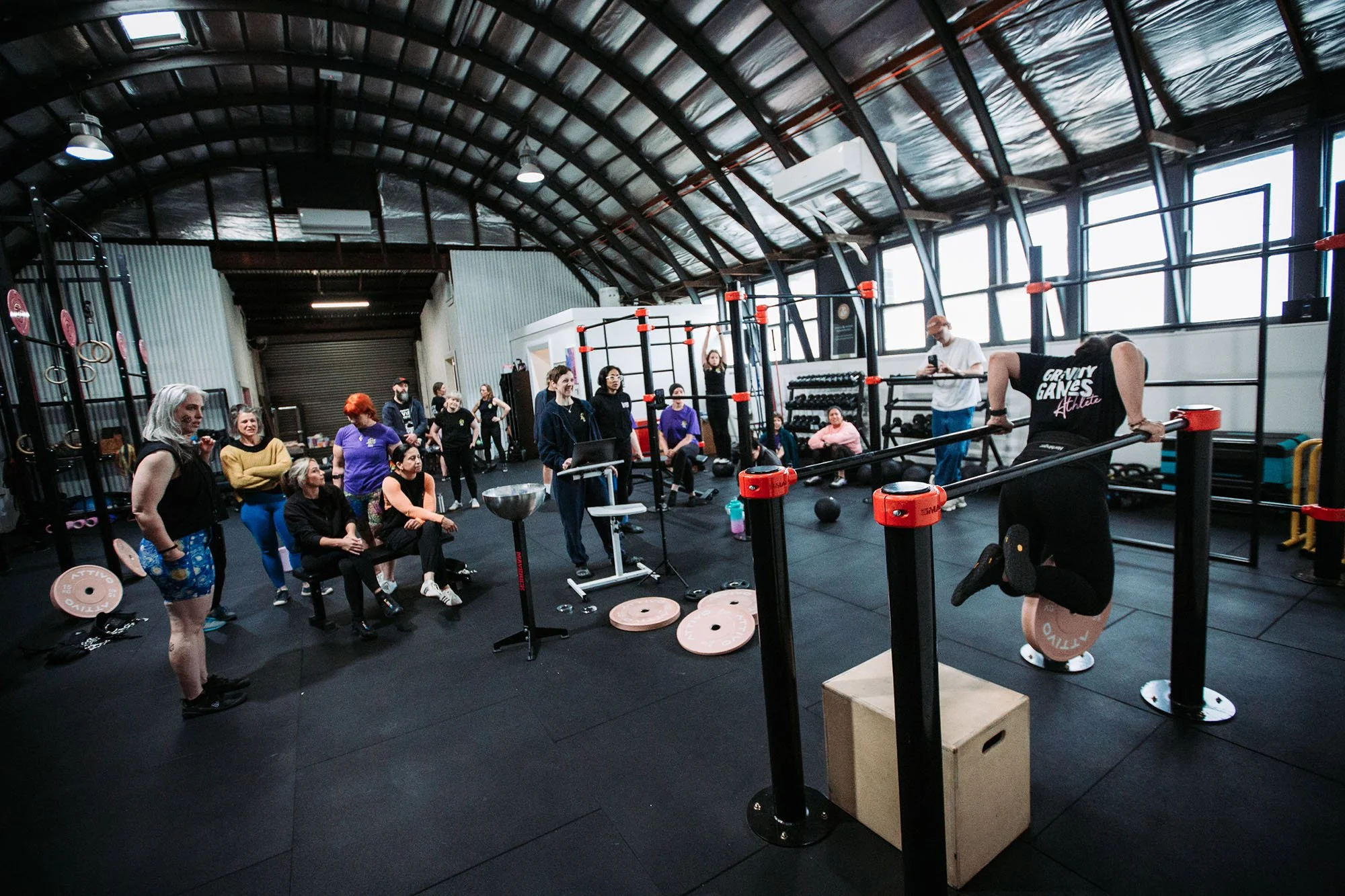 People gathered in a gym for a workout class, with some sitting and others engaging in exercises using gym equipment.