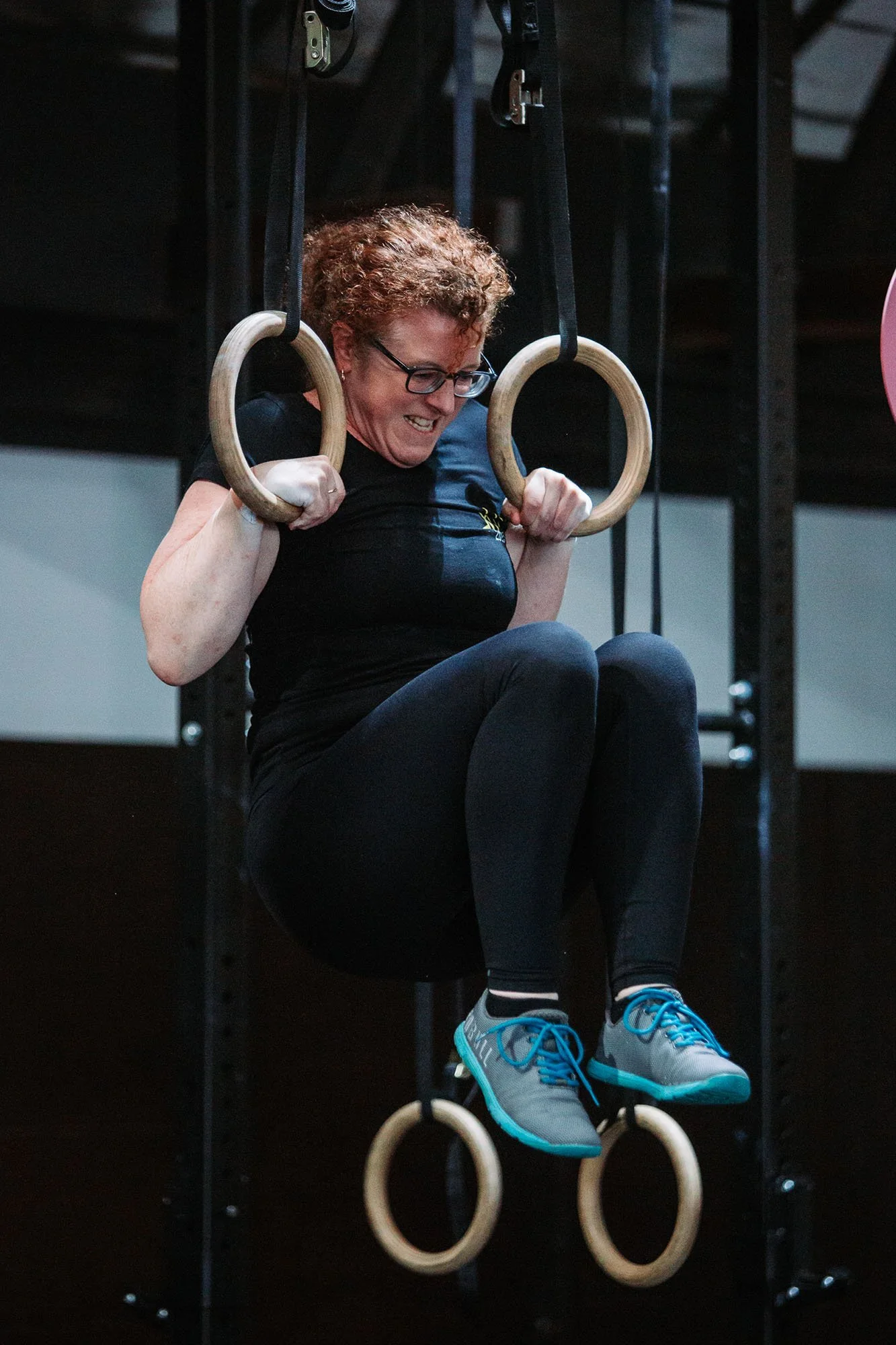 Woman with curly hair and glasses doing a gymnastic exercise on gymnastic rings, wearing a black shirt and gray sneakers with blue accents.