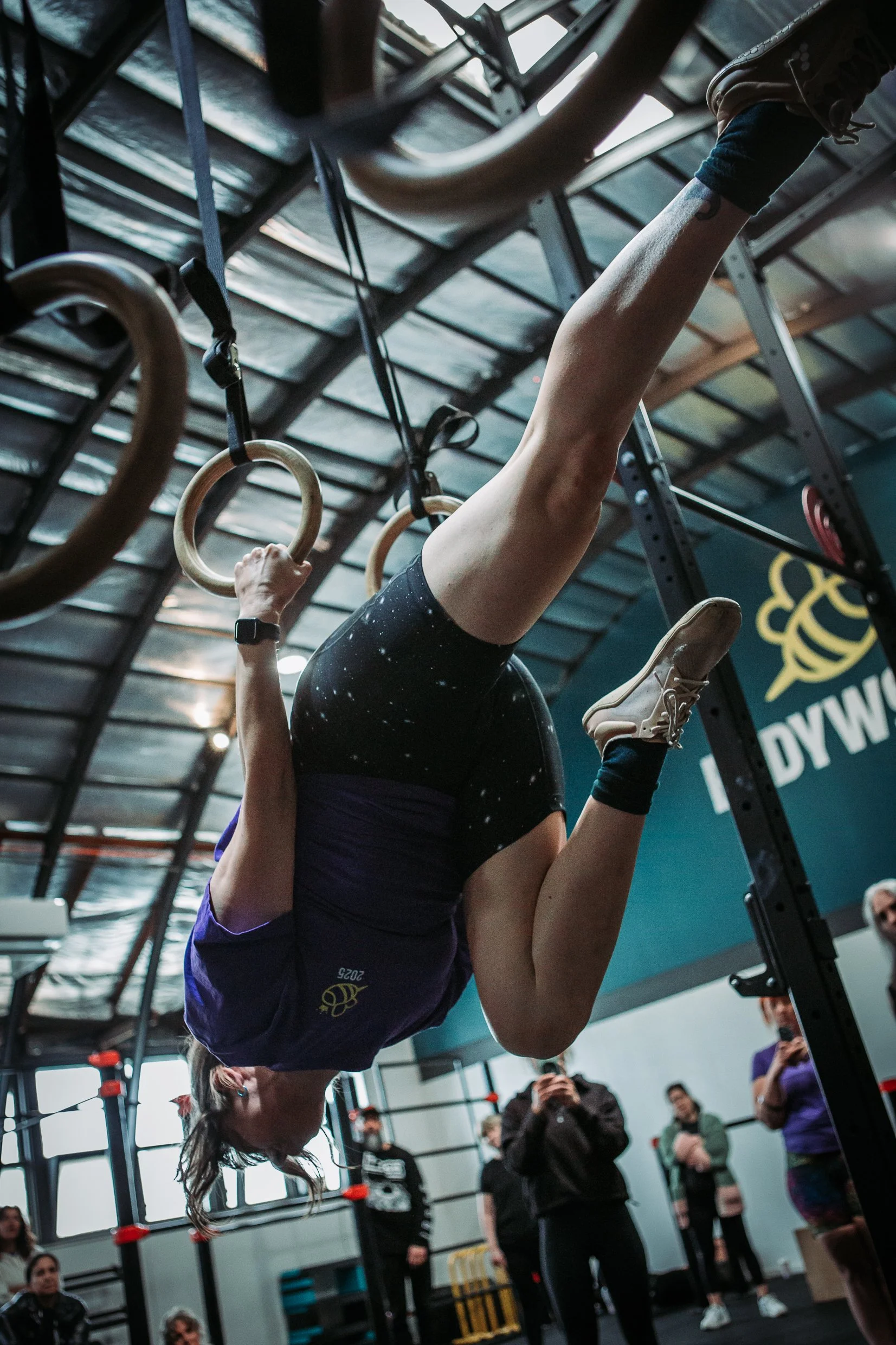 Person hanging upside down and gripping gymnastic rings during workout at gym, surrounded by spectators.