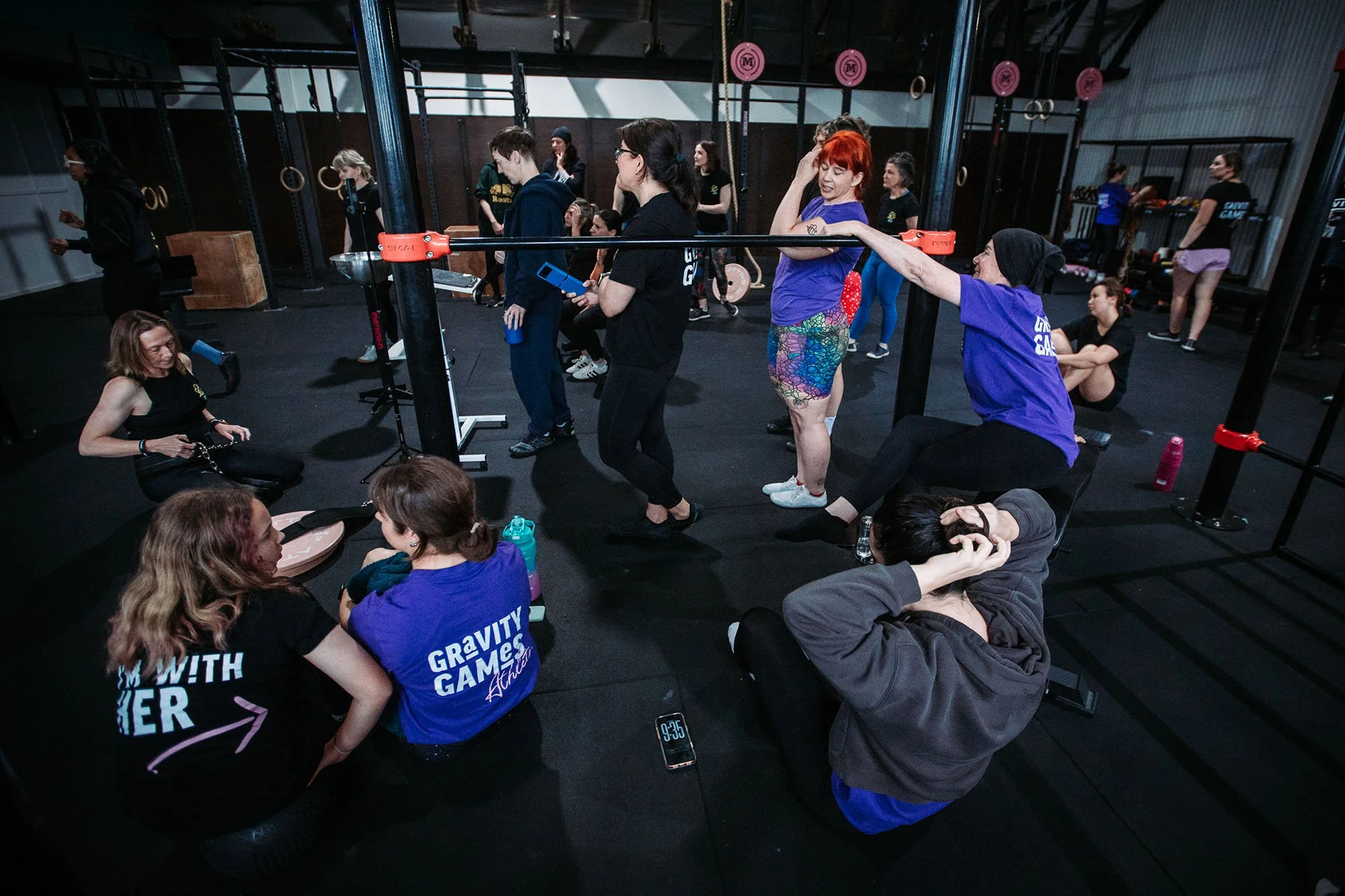 People in a gym participating in a fitness class, with some stretching and some waiting, under gym equipment including rings and barbells.