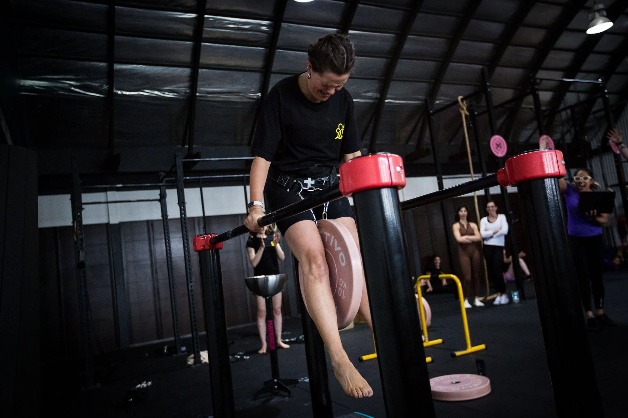 A woman on parallel bars in a gym. Several women are standing and watching in the background, with gym equipment and a black ceiling structure visible.