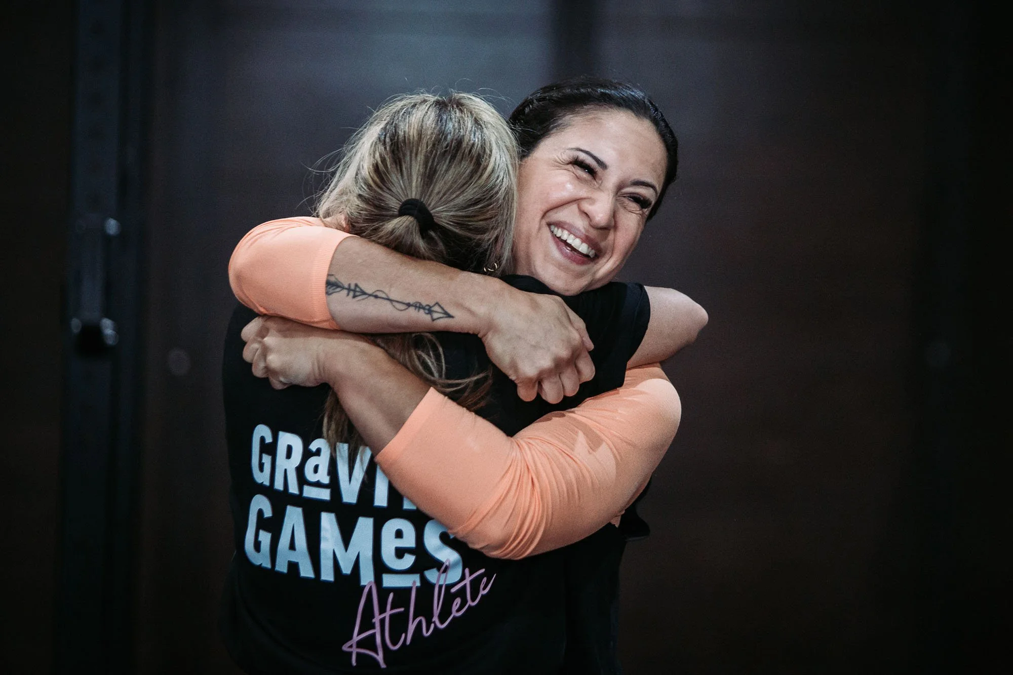 Two women hugging, one with dark hair and the other with blonde hair in a ponytail. The woman with dark hair is smiling. The woman with blonde hair is wearing a black T-shirt with white and pink text, and the woman with dark hair is wearing an orange long-sleeve shirt.