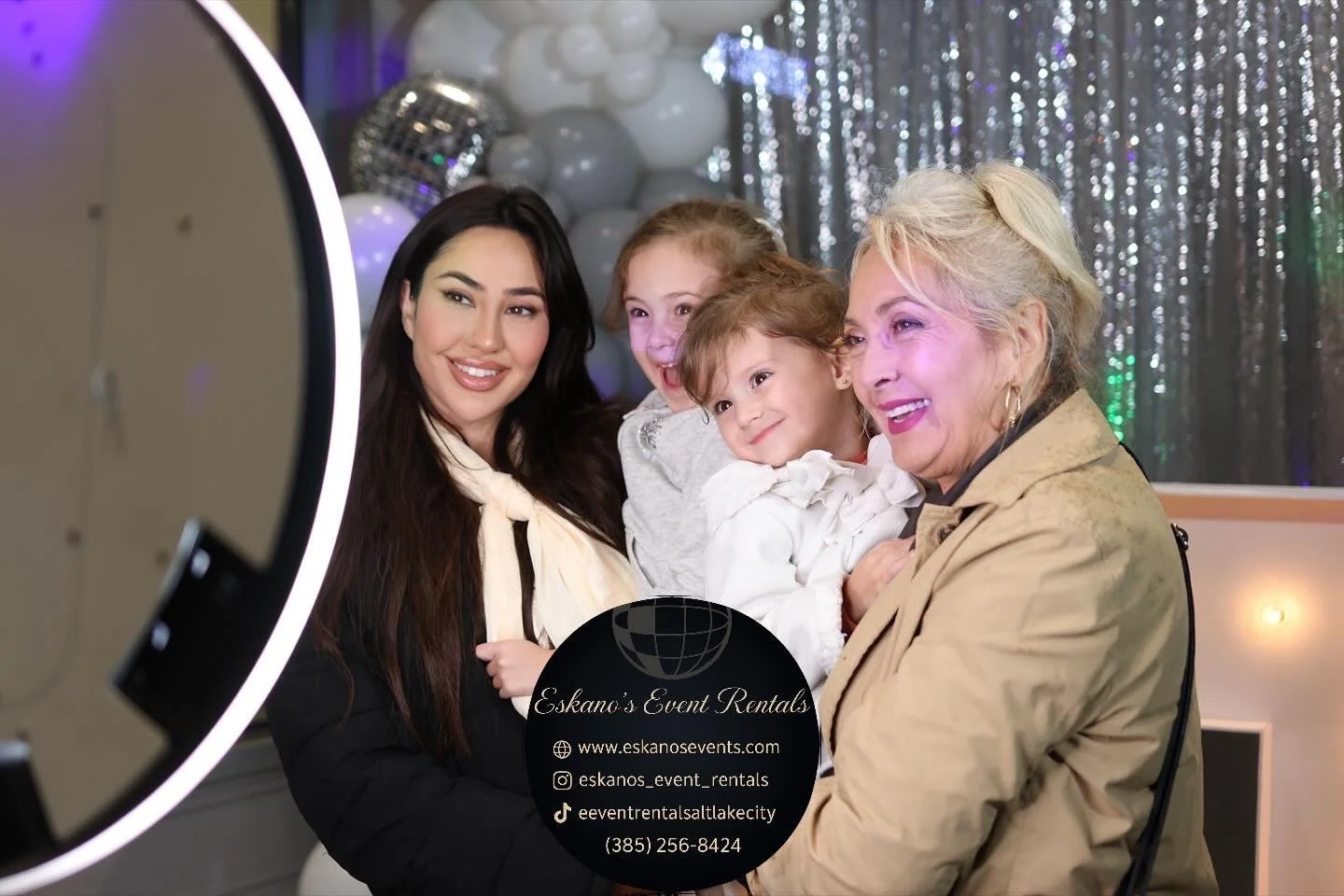 Four women, an older woman and three younger women, are taking a group photo at an event. They are smiling and posing happily, with balloons and a shiny silver curtain in the background. A ring light is partially visible on the left side of the image